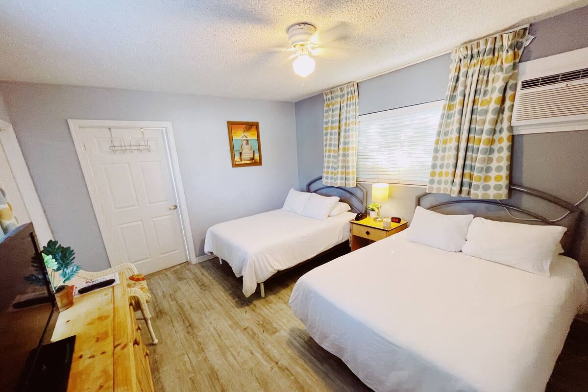 A hotel room with two single beds, a wooden desk, a chair, a nightstand with a lamp, a painting on the wall, and a window with curtains.