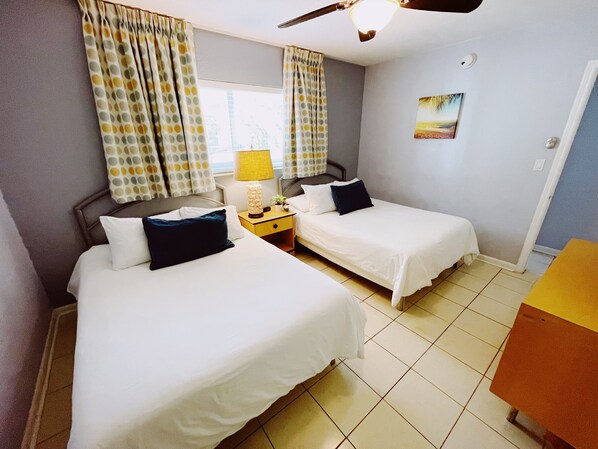 A hotel room with two beds, a nightstand with a lamp, a ceiling fan, and a painting on the wall.