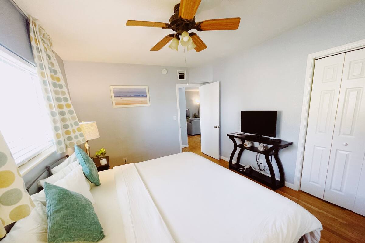 A bedroom with a bed, a ceiling fan, a TV on a stand, and a door leading to another room.