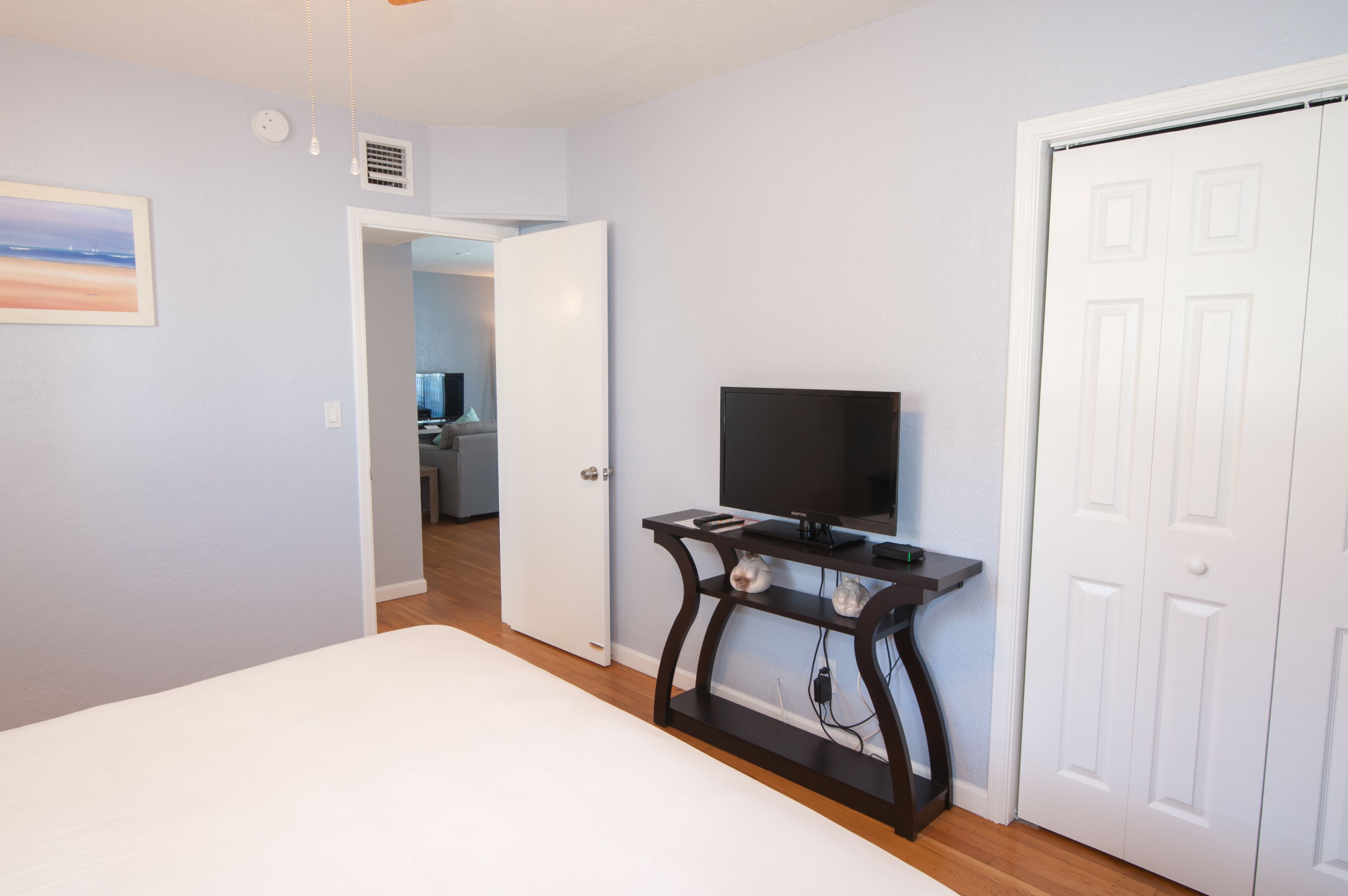 A bedroom with a bed, a TV on a black stand, a white door, and a hallway leading to a bathroom.