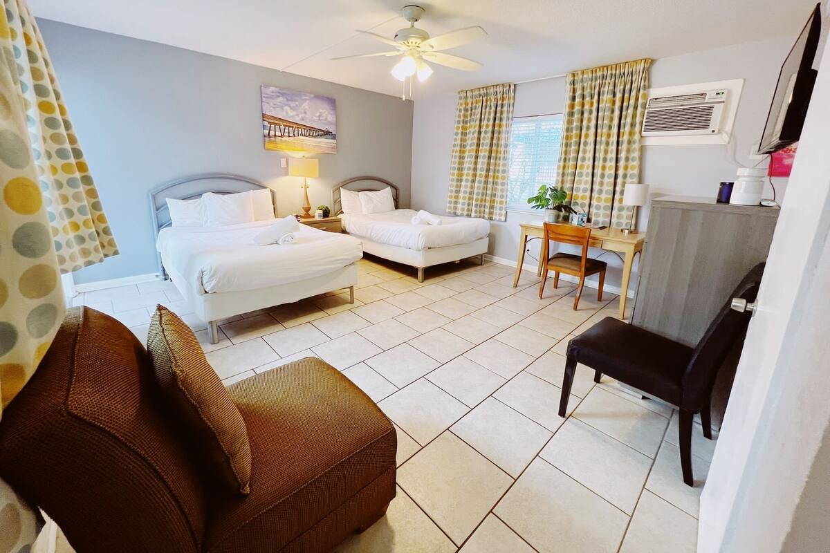 A hotel room with two beds, a desk, a chair, a ceiling fan, and a window with curtains.