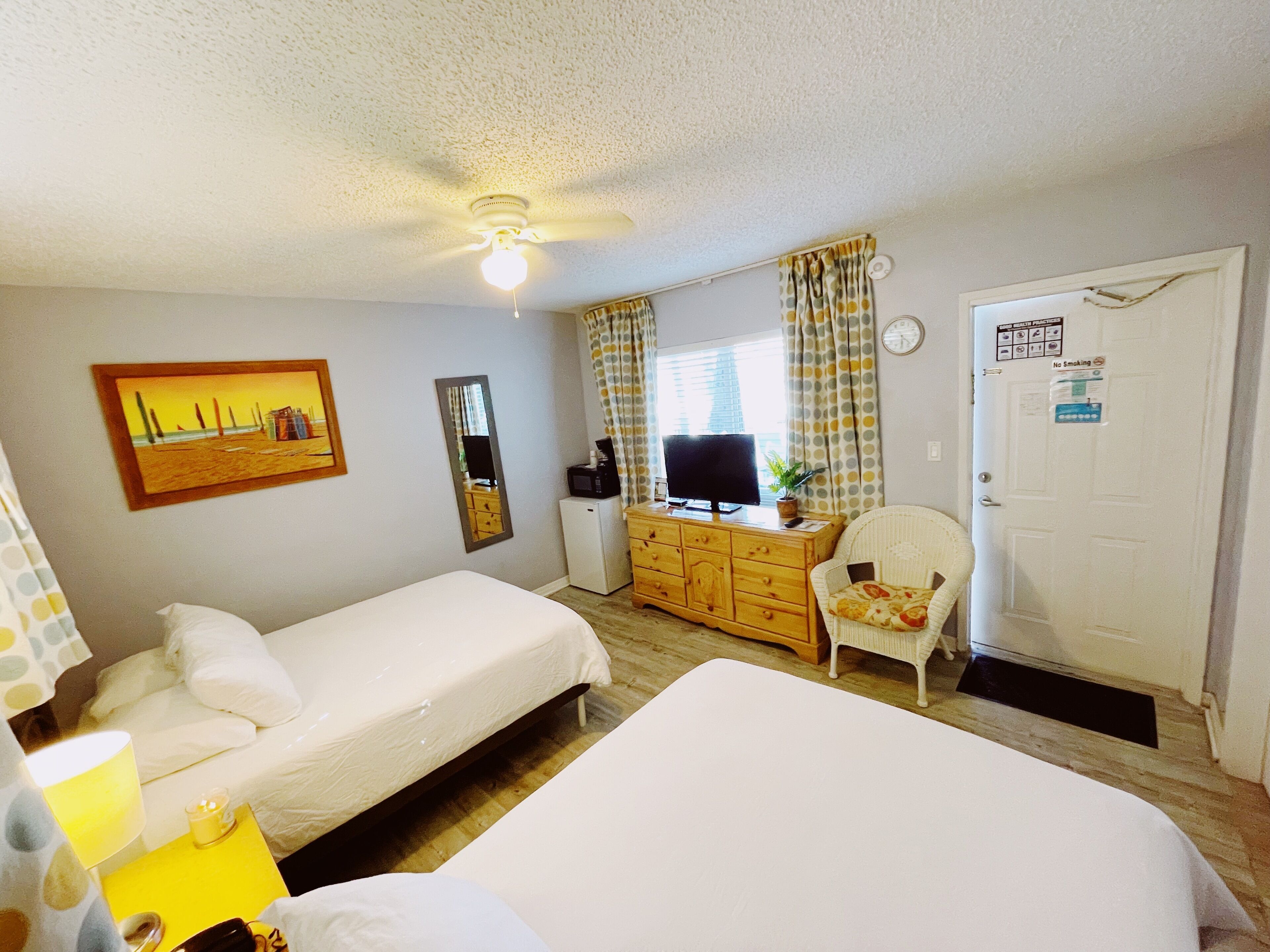 A hotel room with two beds, a desk with a television, a chair, a dresser, a ceiling fan, and a painting on the wall.