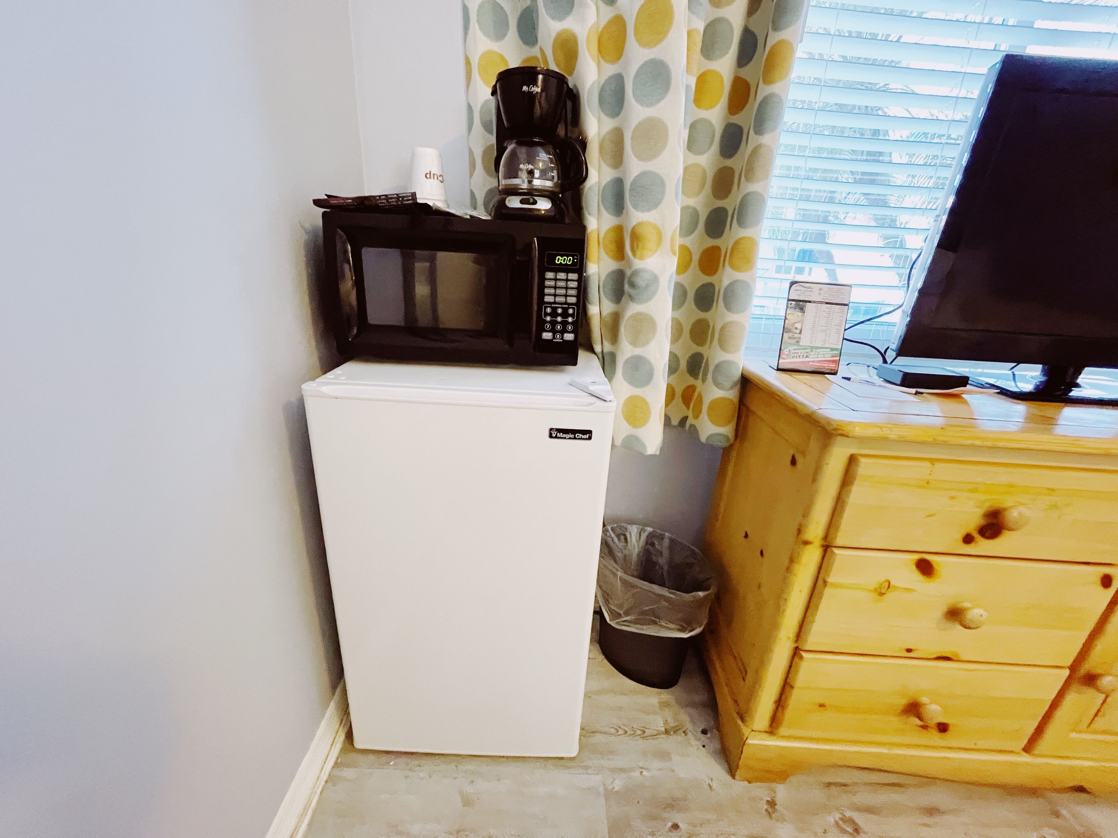 Een compacte kitchenette met een witte koelkast, een zwarte magnetron, en een koffiezetapparaat erboven. Een houten kast met lades bevindt zich rechts, en een prullenbak staat onder de magnetron.