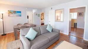 A living room with a gray sofa, a dining table with chairs, and a painting of a beach scene. - Great Escape Apartments by Lowkl (Lauderdale-by-the-Sea)