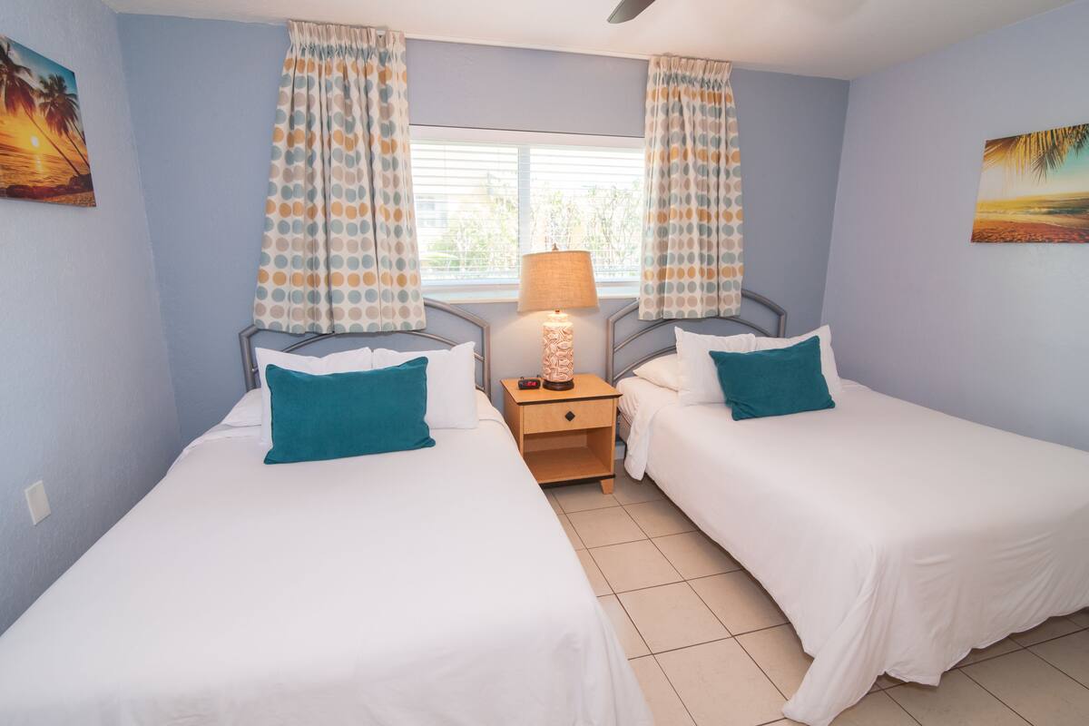 A room with two single beds, a nightstand with a lamp, and a window with curtains.