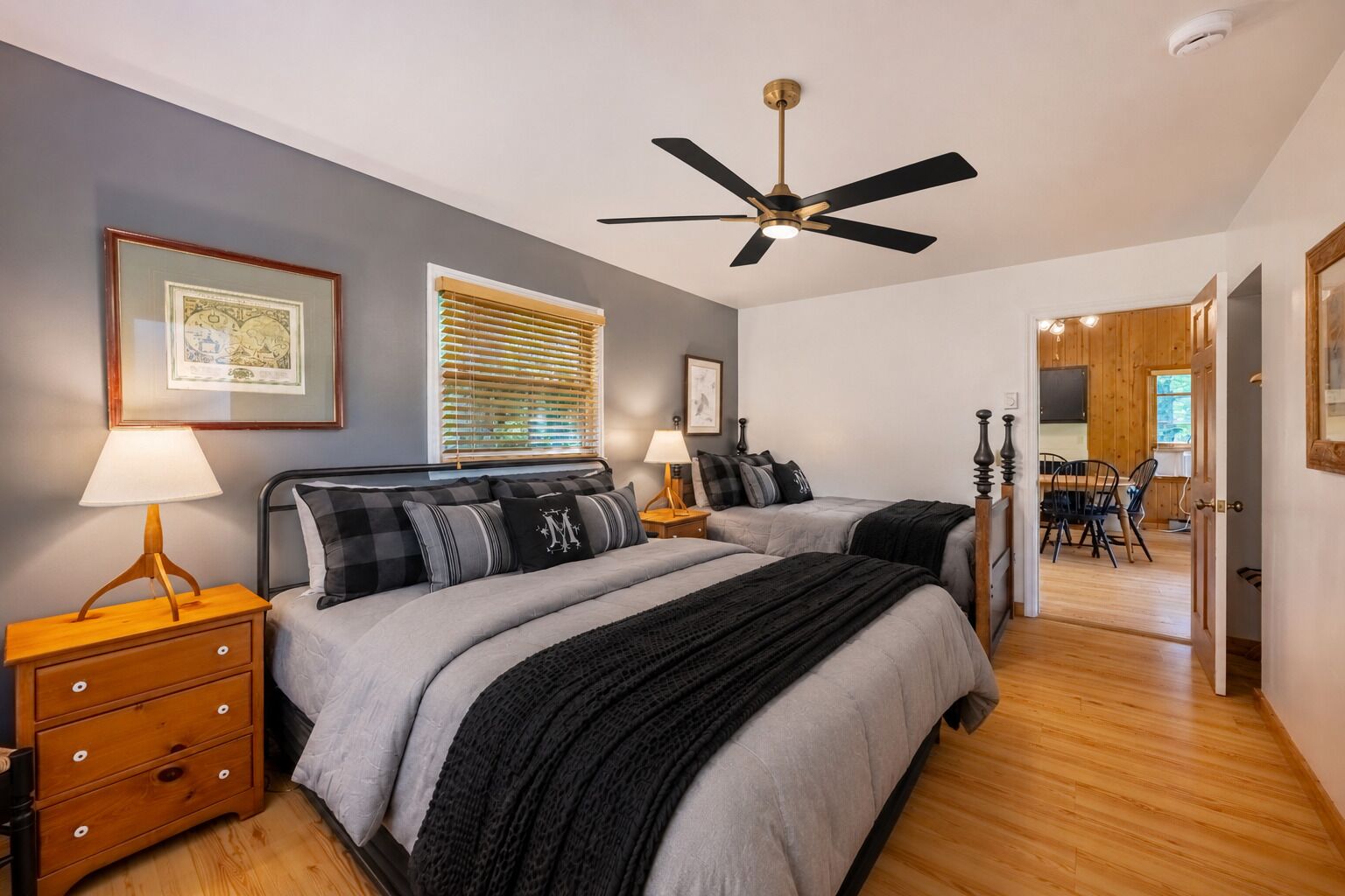 A bedroom with two beds, a ceiling fan, wooden flooring, and a dining area visible through an open door.