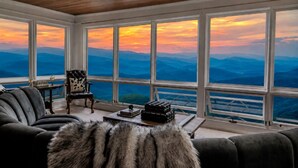 Una stanza con una grande finestra vista montagna, una poltrona in pelle, una coperta di pelliccia e un tavolino con un libro e un bicchiere.