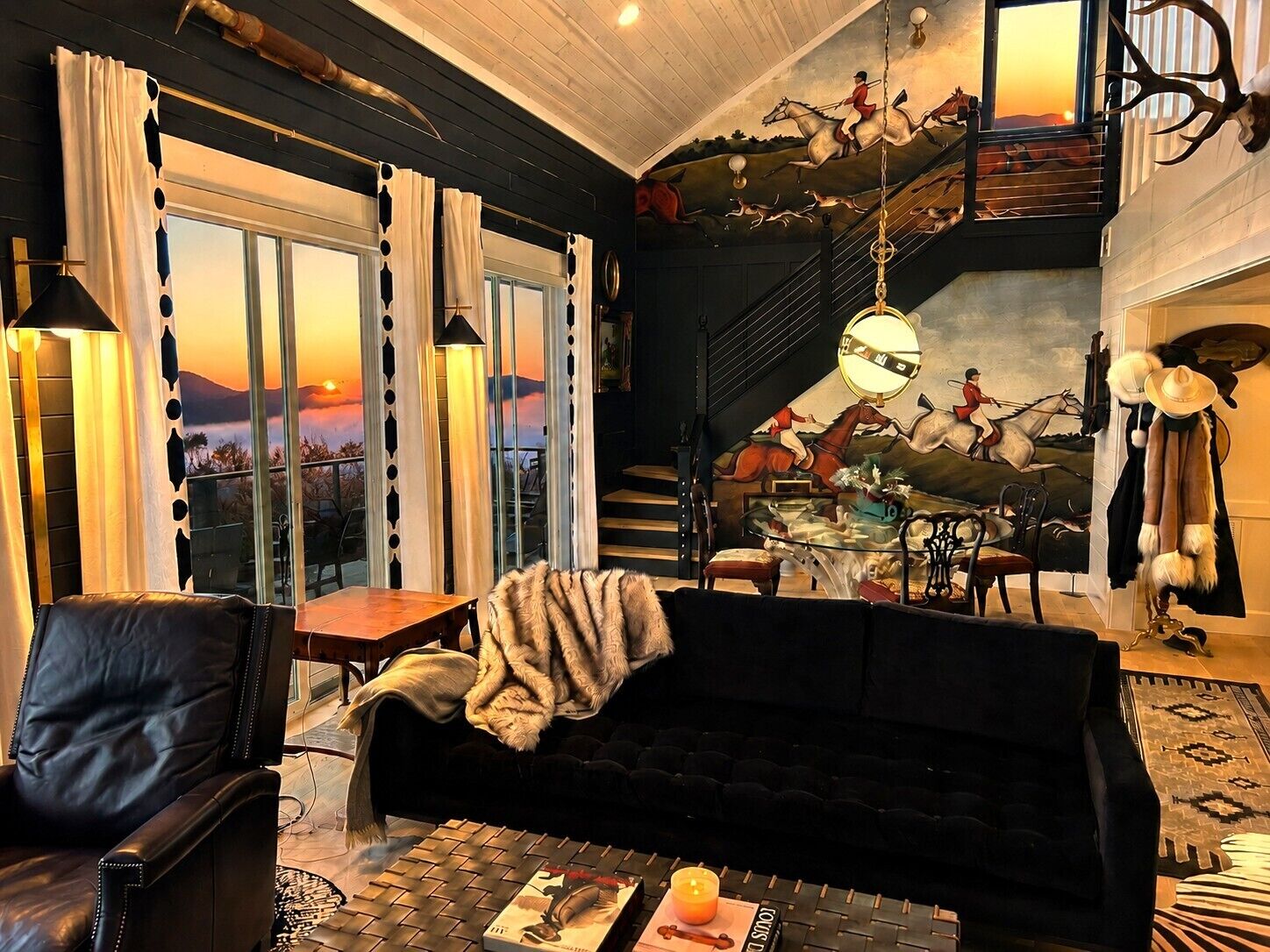 A cozy living room with a leather sofa, a coffee table with a book and candle, and a scenic view through large windows.