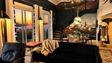 A cozy living room with a leather sofa, a coffee table with a book and candle, and a scenic view through large windows.