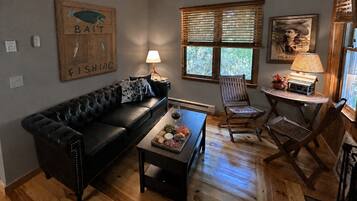 A cozy living room with a black leather sofa, a wooden coffee table with snacks, a lamp, and a window with blinds.