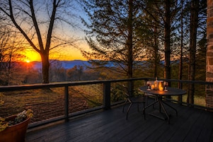 A wooden deck with a table and chairs, overlooking a forested landscape with a sunset.