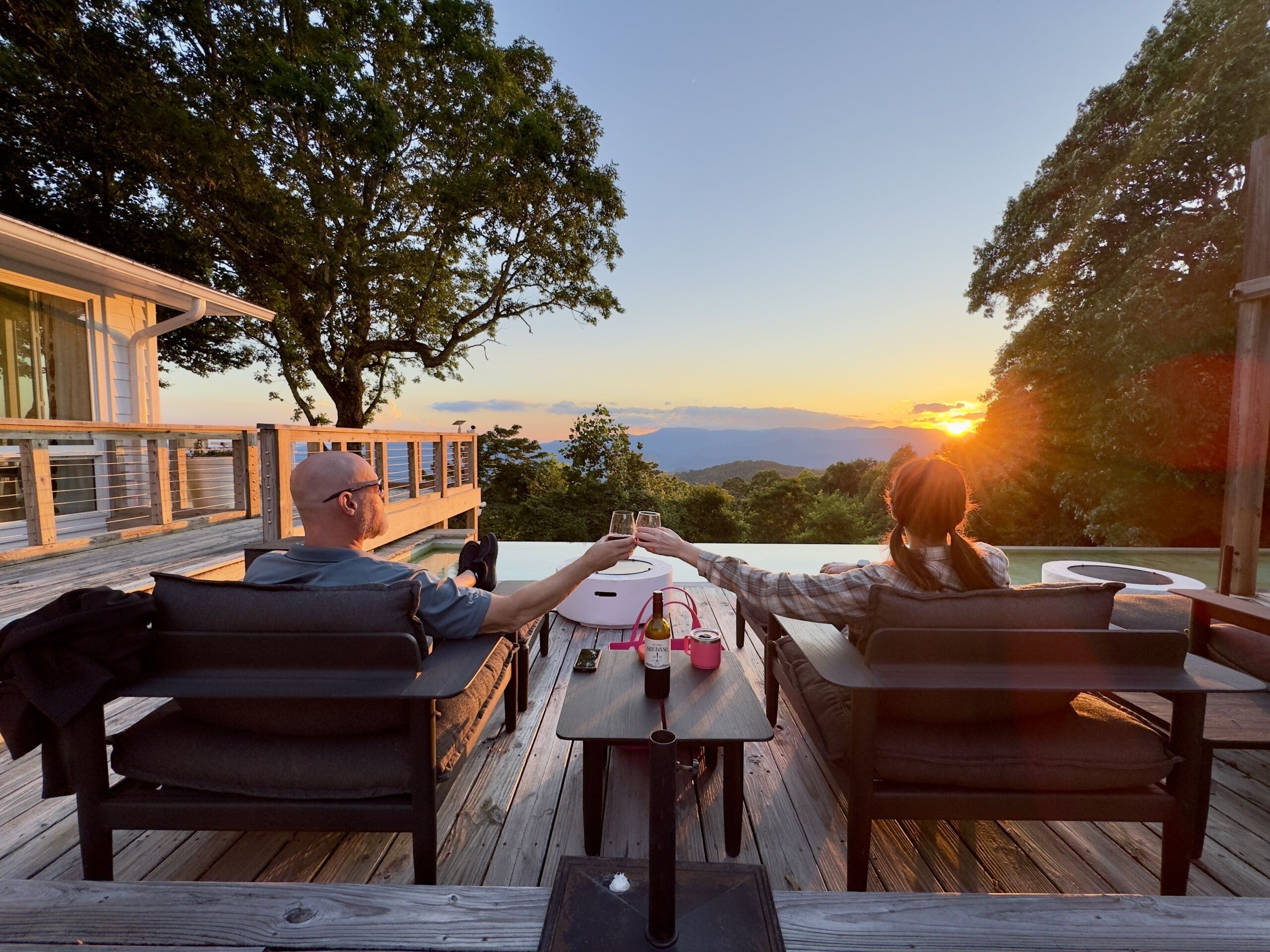 Two people seated at a wooden deck table with a sunset view, a bottle of wine, and a glass.