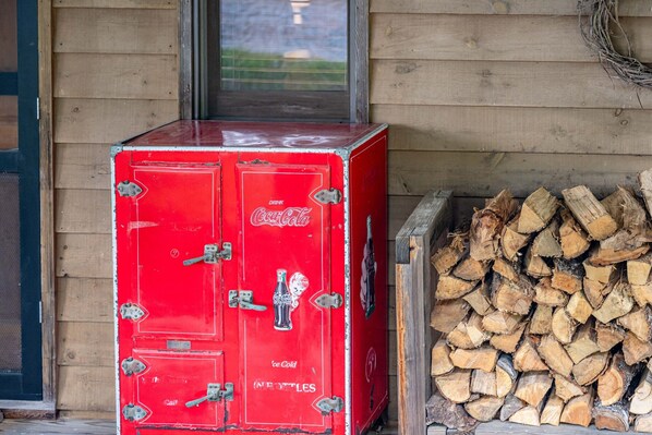 A red Coca-Cola ice cooler with a Coca-Cola logo, placed next to a stack of firewood.