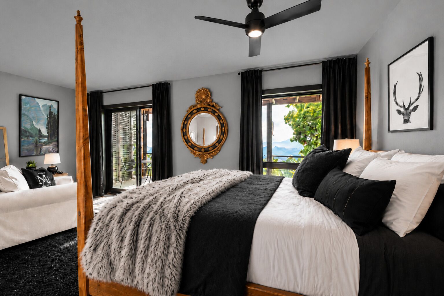 A bedroom with a large bed, a ceiling fan, a window with a view, and a mirror on the wall.