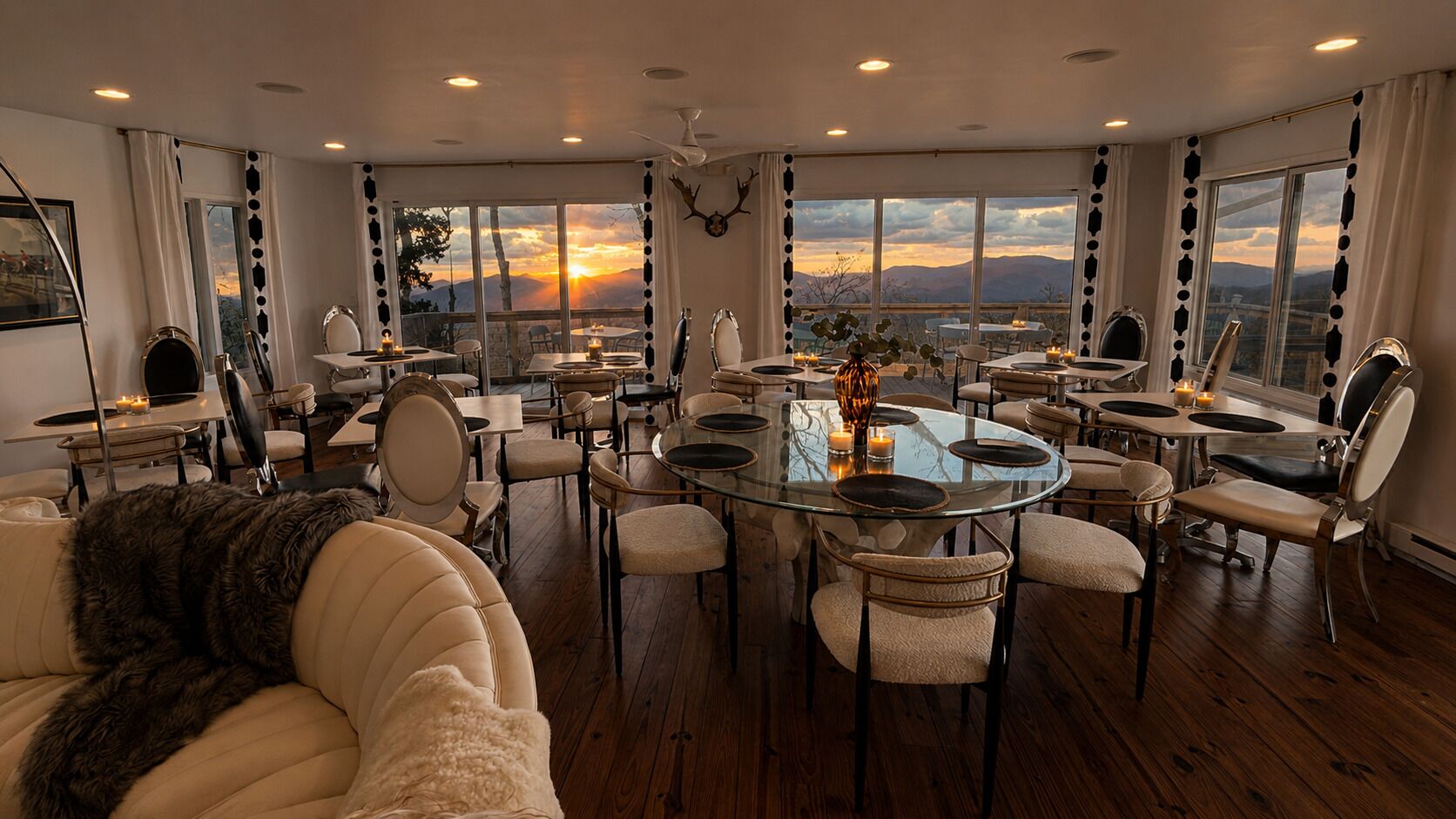 A spacious dining area with round tables and chairs, large windows with a sunset view, and a cozy seating area with a fur throw.