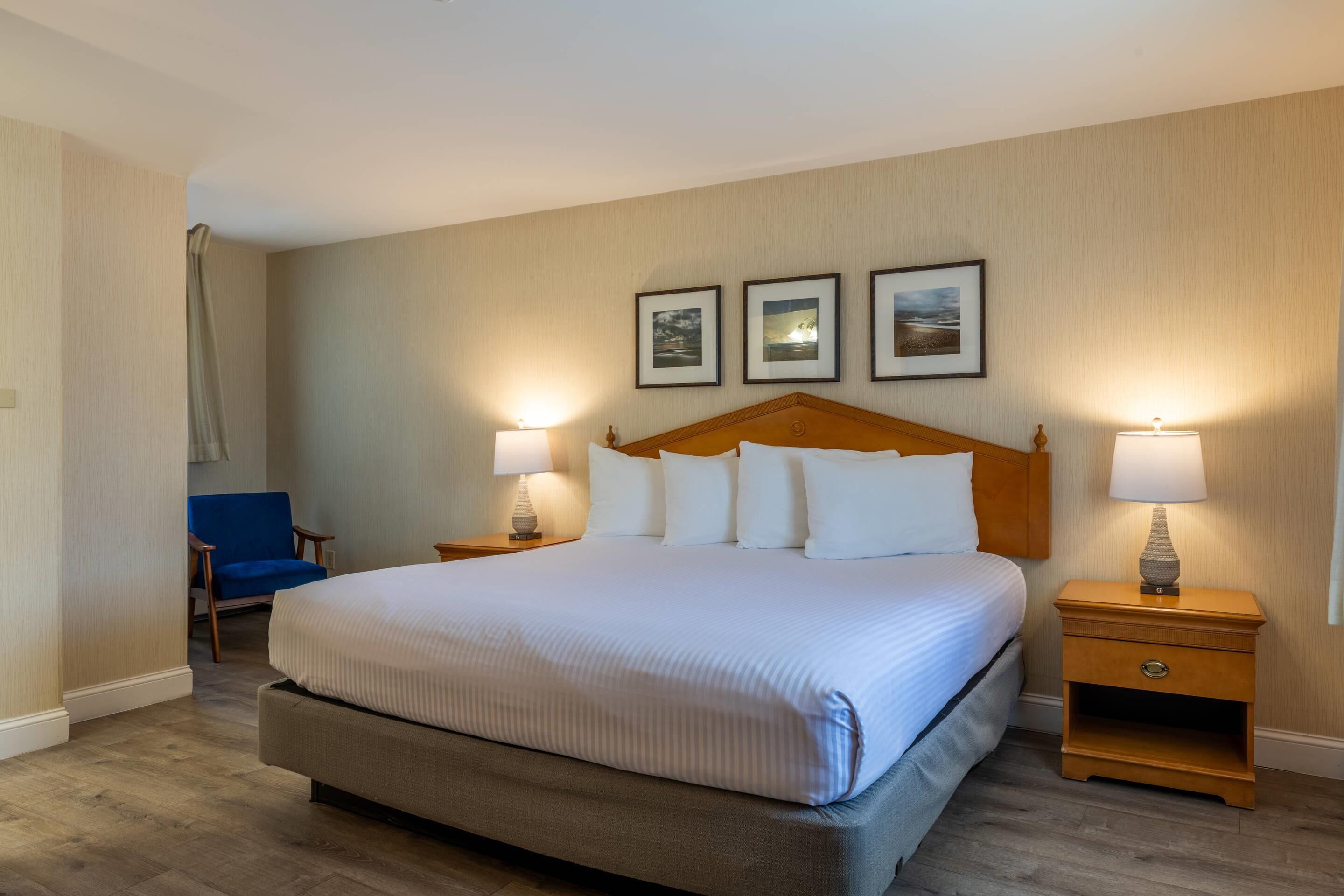 Habitación de hotel con una cama grande, dos mesitas de noche con lámparas, una silla y tres cuadros en la pared.