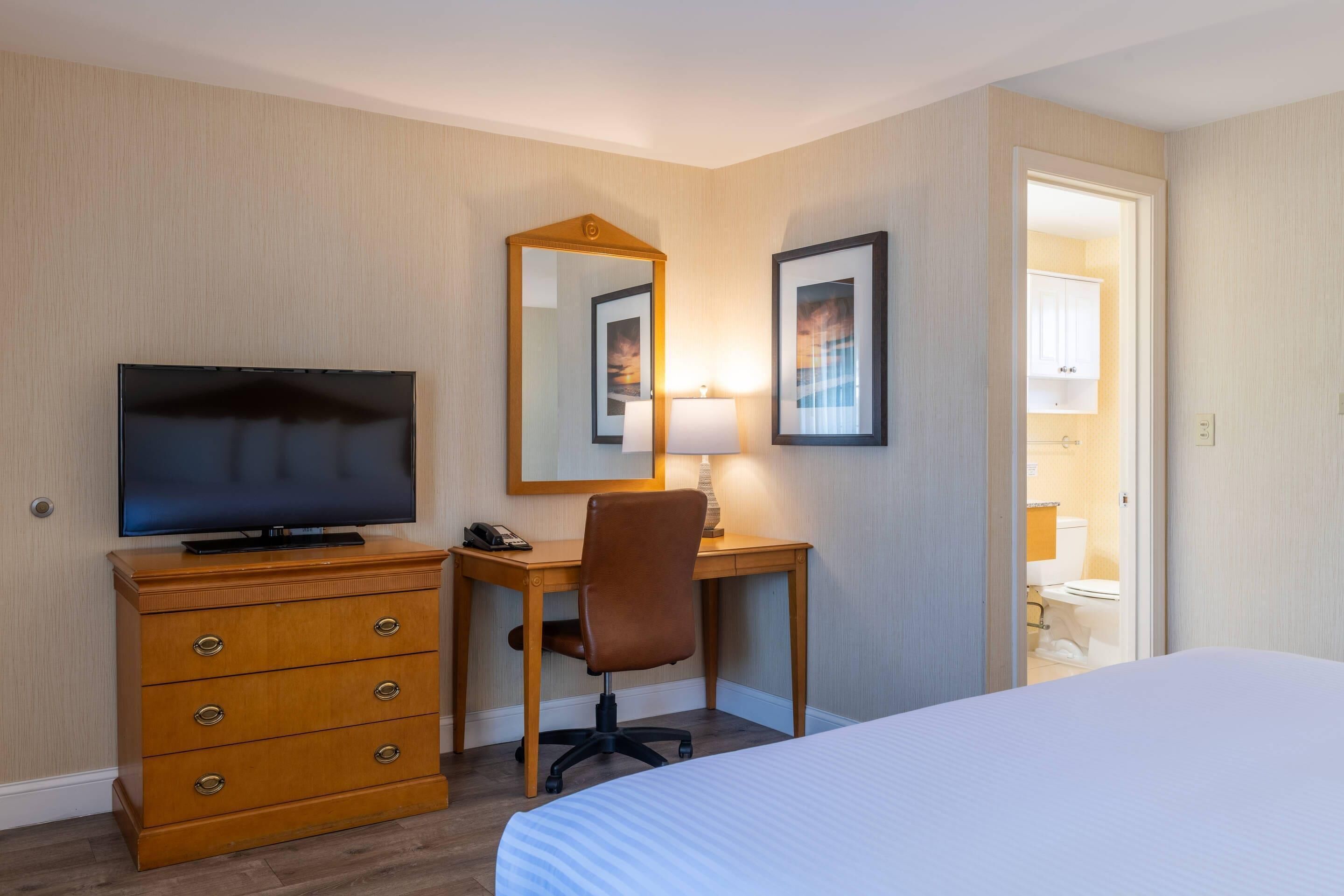 Habitación de hotel con cama, escritorio con silla, televisor, cómoda y baño visible a través de una puerta abierta.