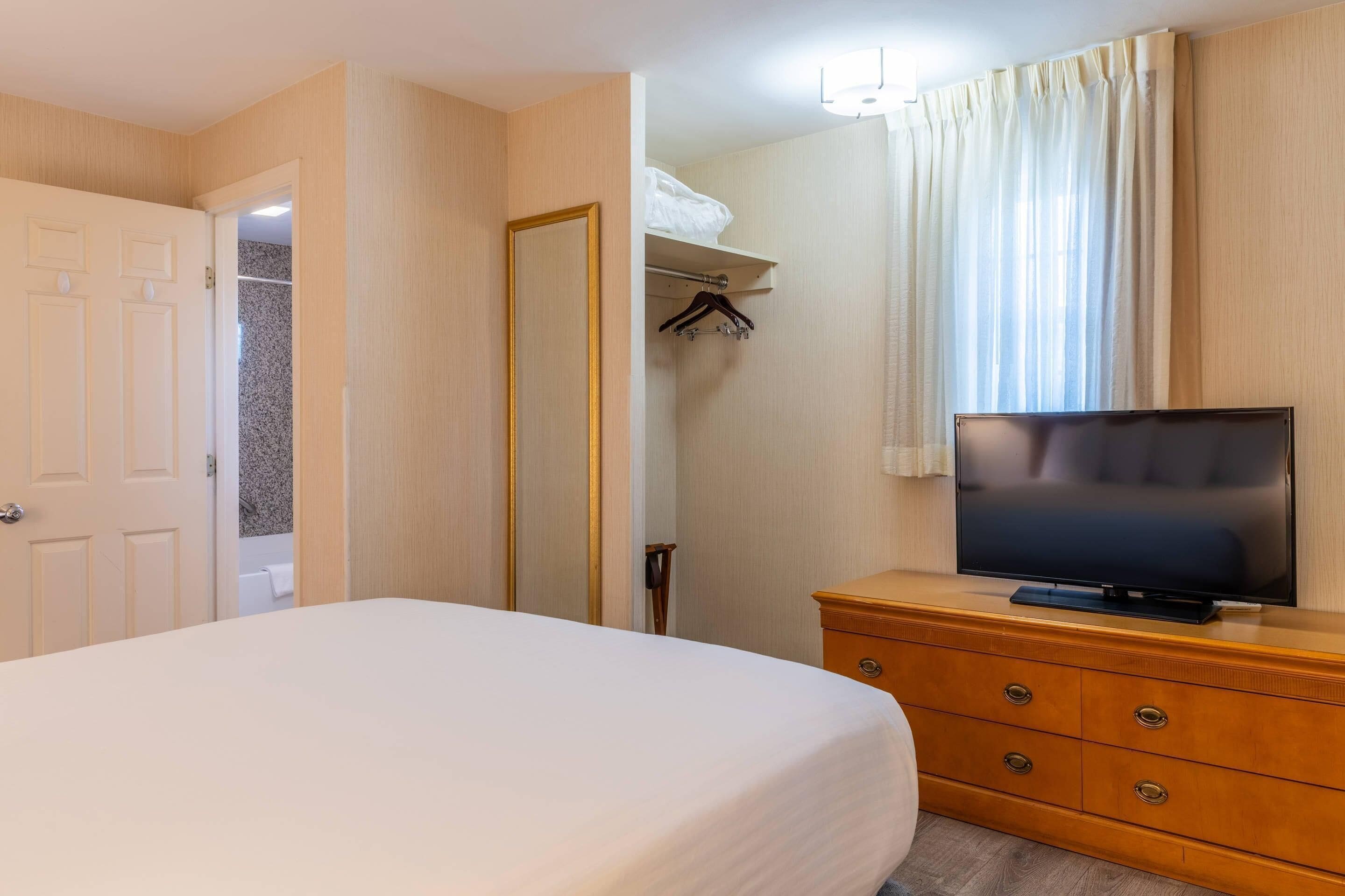 Habitación de hotel con cama, cómoda de madera, televisor, armario y baño visible a través de una puerta abierta.
