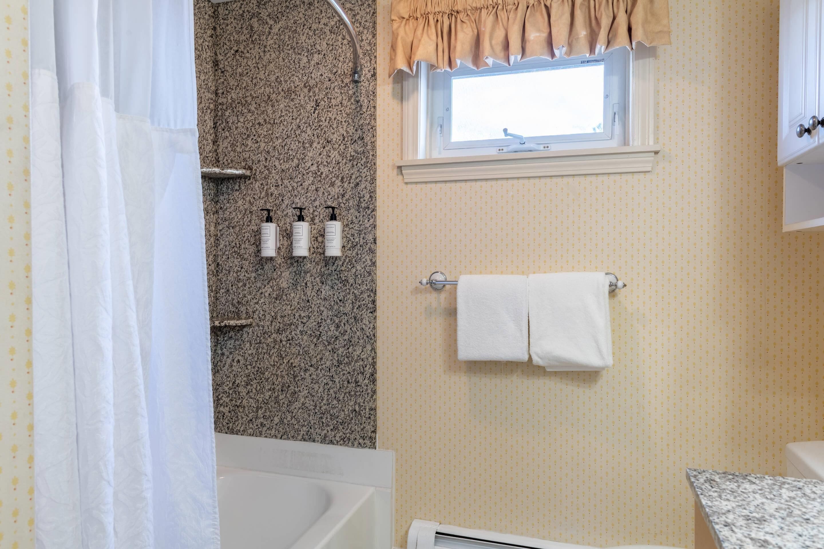 A bathroom with a bathtub, a window with a curtain, a towel rack with towels, and toiletries on the wall.