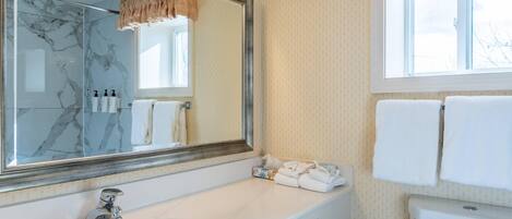 A bathroom with a marble-tiled shower, a large mirror, a white sink, and a toilet.