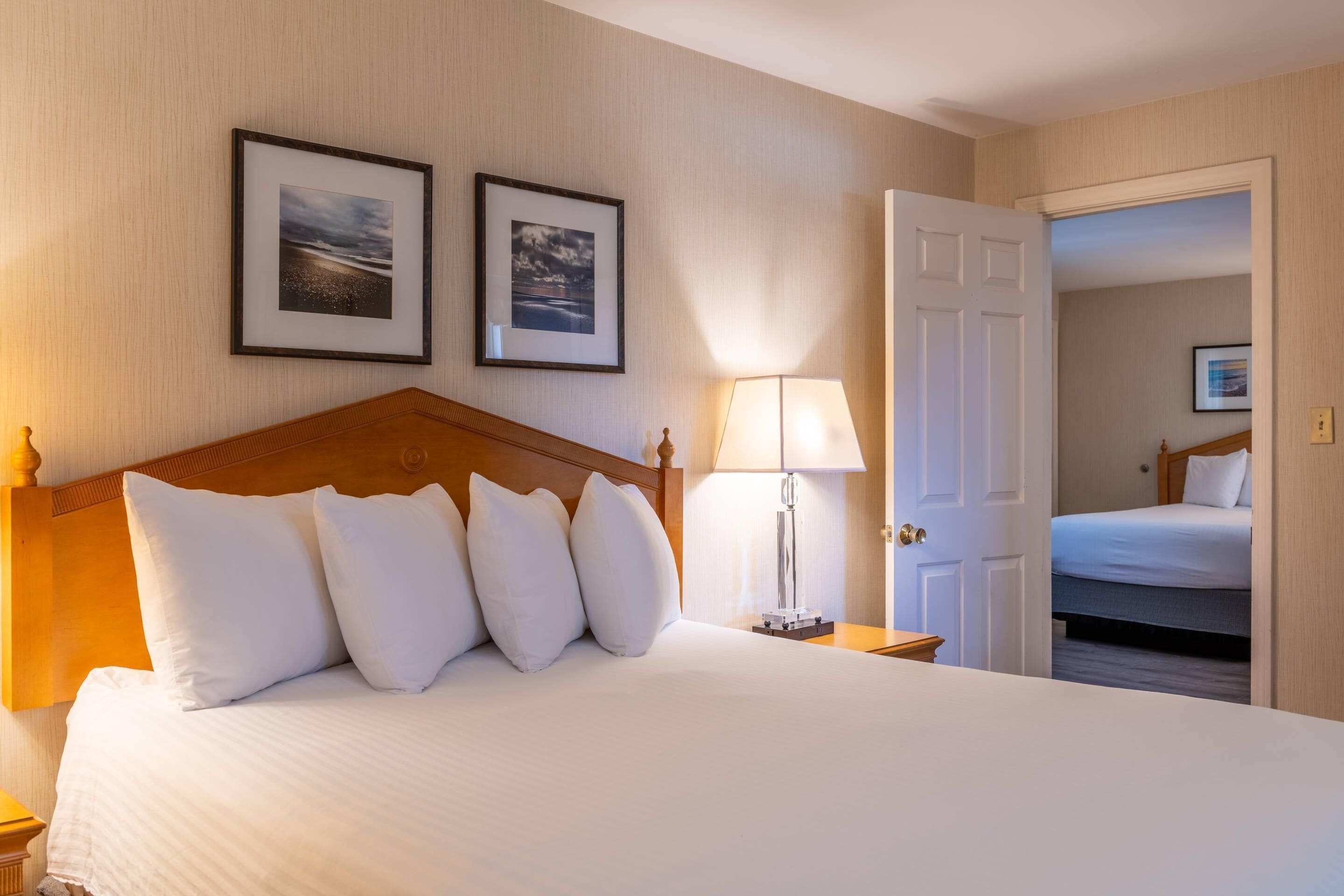 Habitació d'hotel amb un llit gran, una tauleta de nit amb una làmpada, dues imatges emmarcades a la paret i una porta oberta que condueix a una altra habitació.