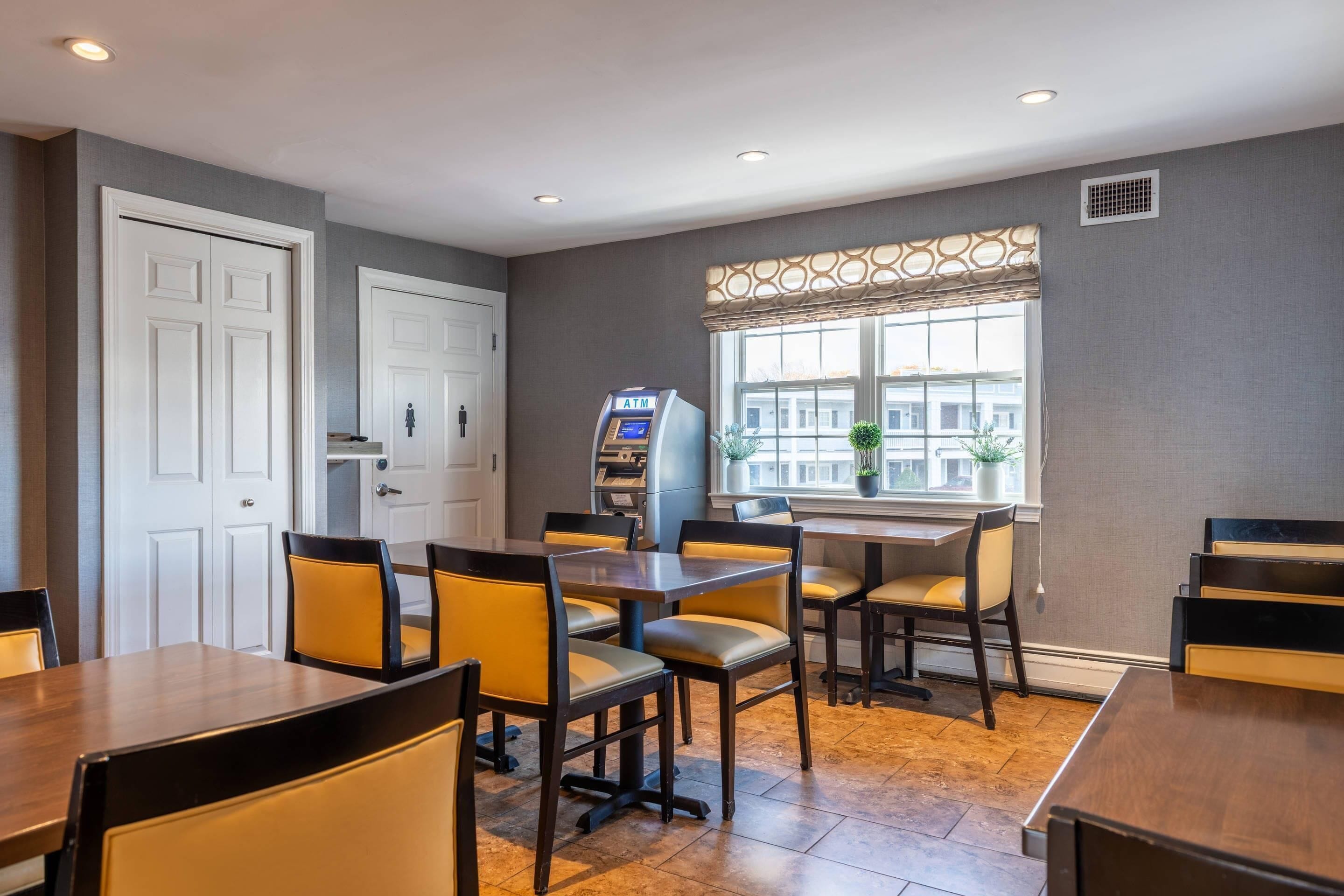 A dining area with tables and chairs, an ATM machine, and a window with a view of buildings.