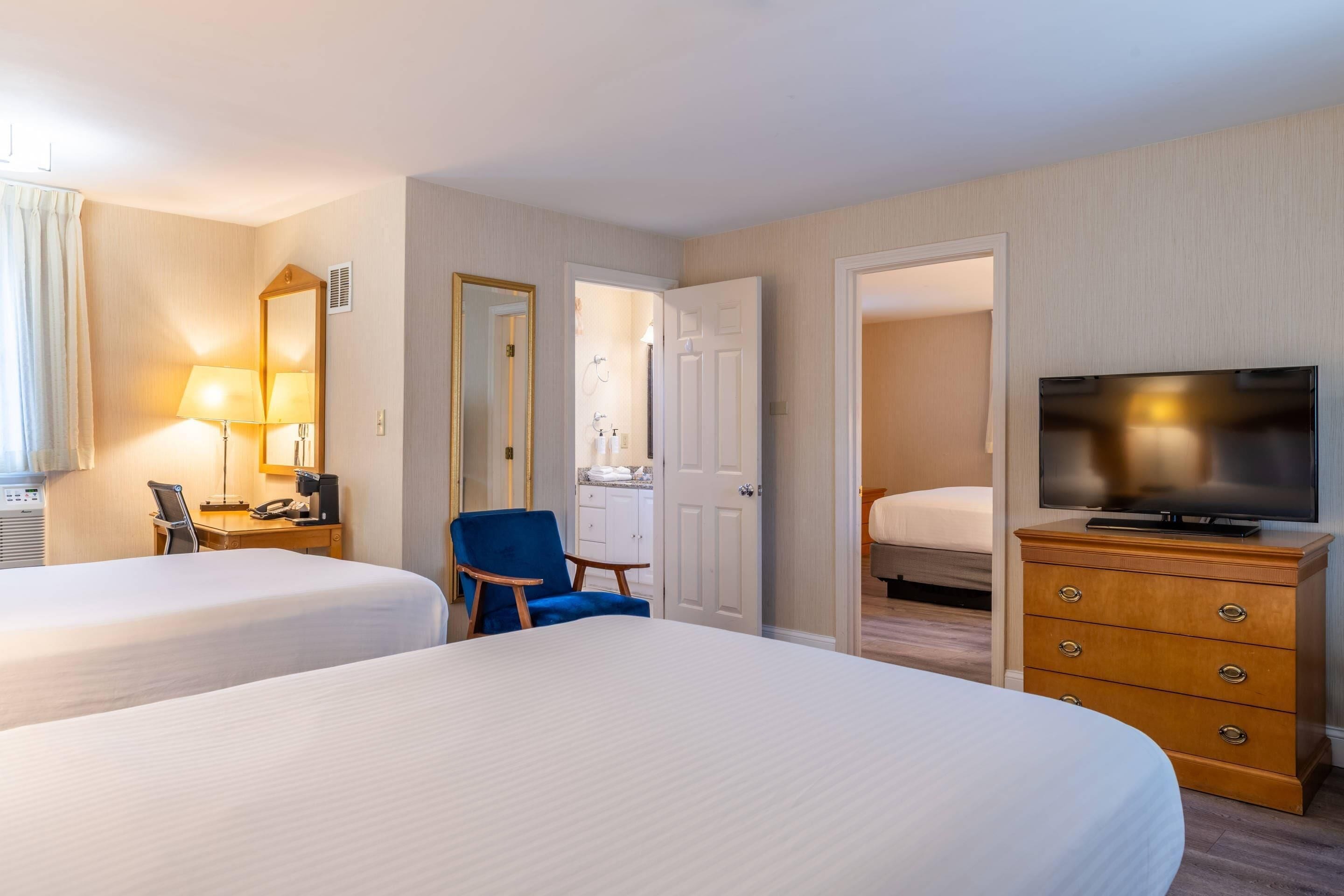 A hotel room with two beds, a desk with a lamp, a chair, a dresser with a TV, and a bathroom visible through an open door.