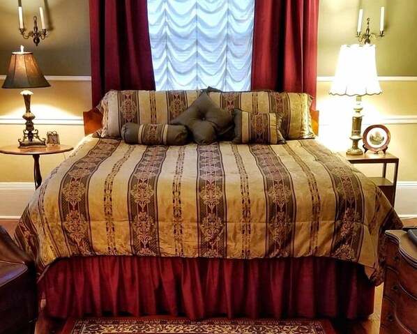 A neatly made bed with a patterned bedspread and pillows, a red curtain, and a lamp on a bedside table.