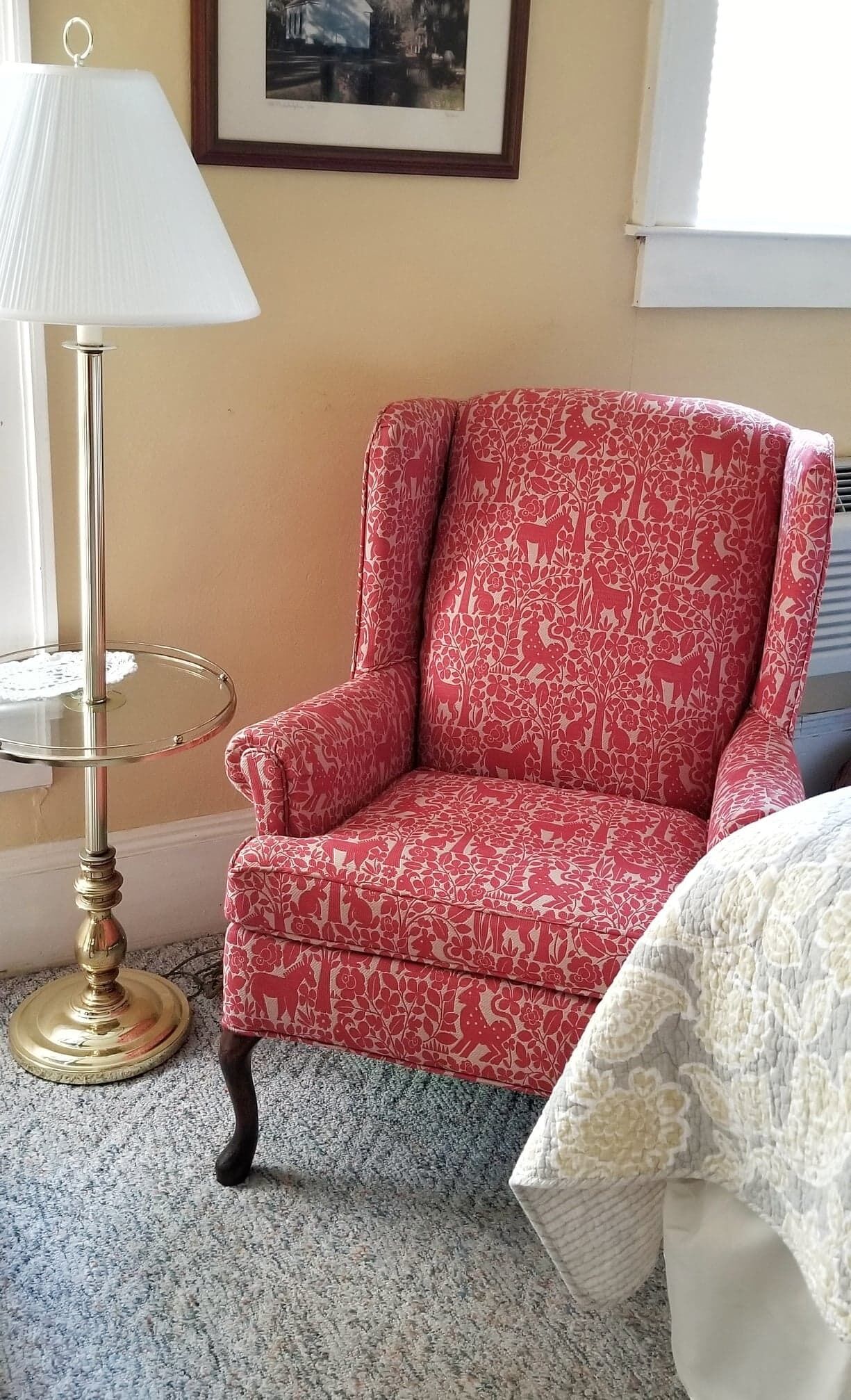 A room with a red patterned armchair, a gold floor lamp, and a framed picture on the wall.