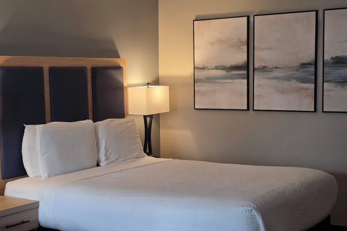A neatly made bed with white linens, a bedside lamp, and three framed artworks on the wall.