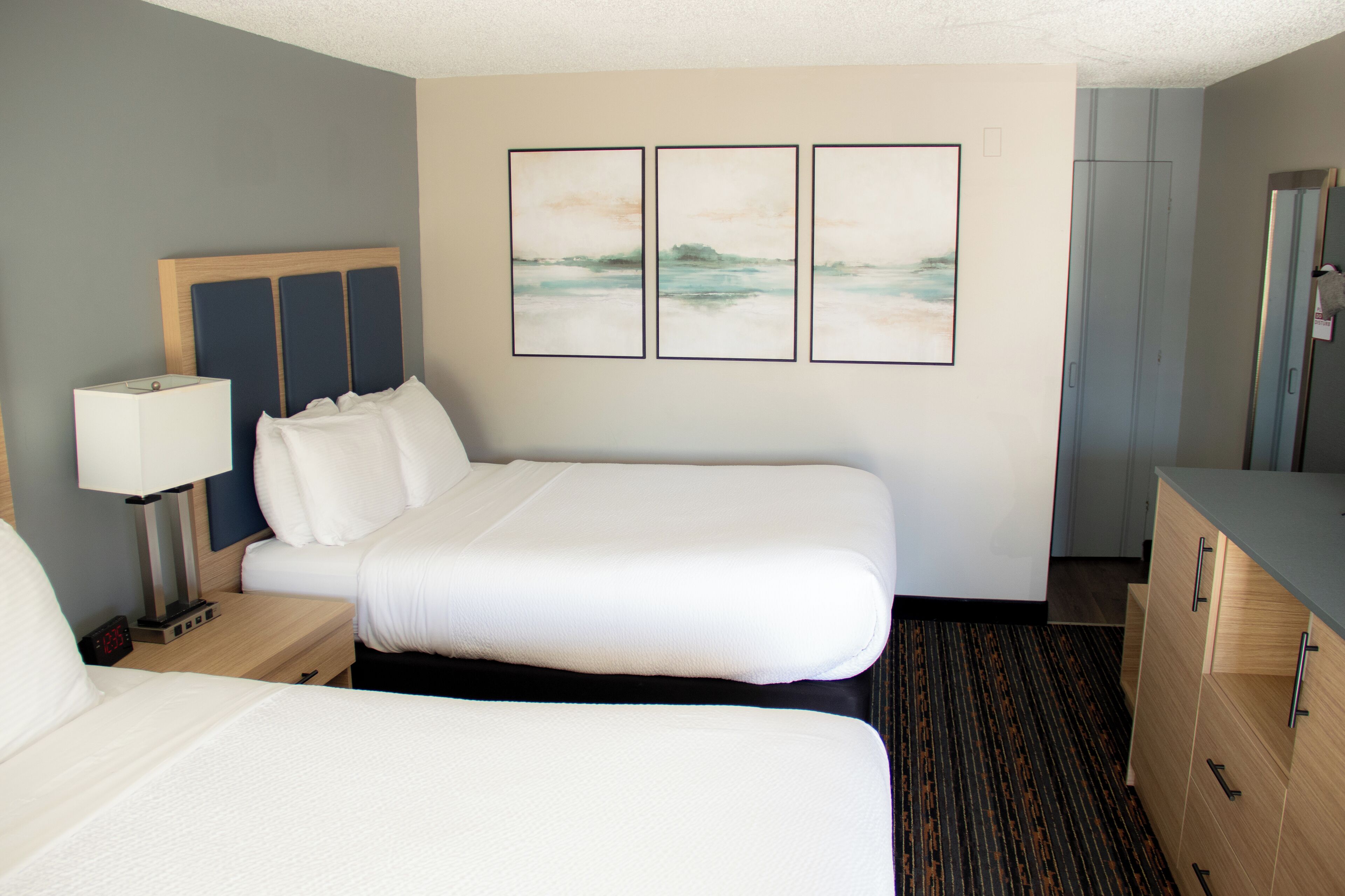 Habitación de hotel con cama doble, mesita de noche con lámpara, cómoda y tres cuadros en la pared.
