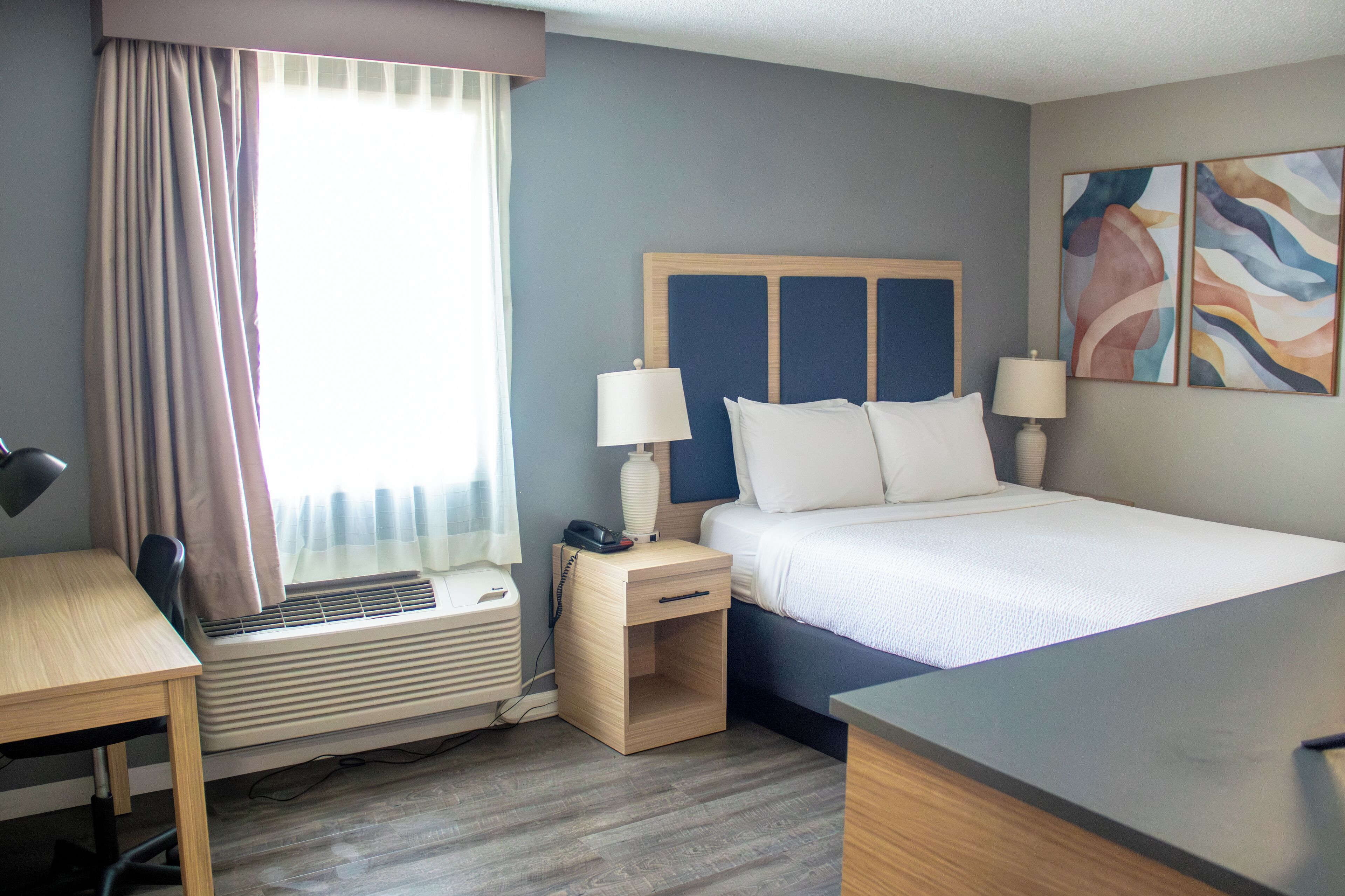 Habitación de hotel con cama, mesitas de noche, escritorio, silla, equipo de aire acondicionado y ventana con cortinas.