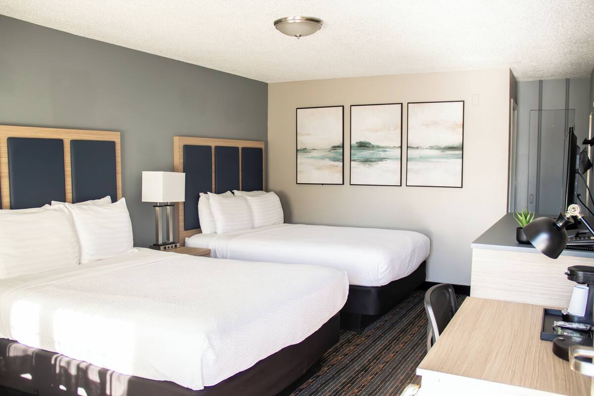 A hotel room with two beds, a desk, a chair, a lamp, and three framed pictures on the wall.