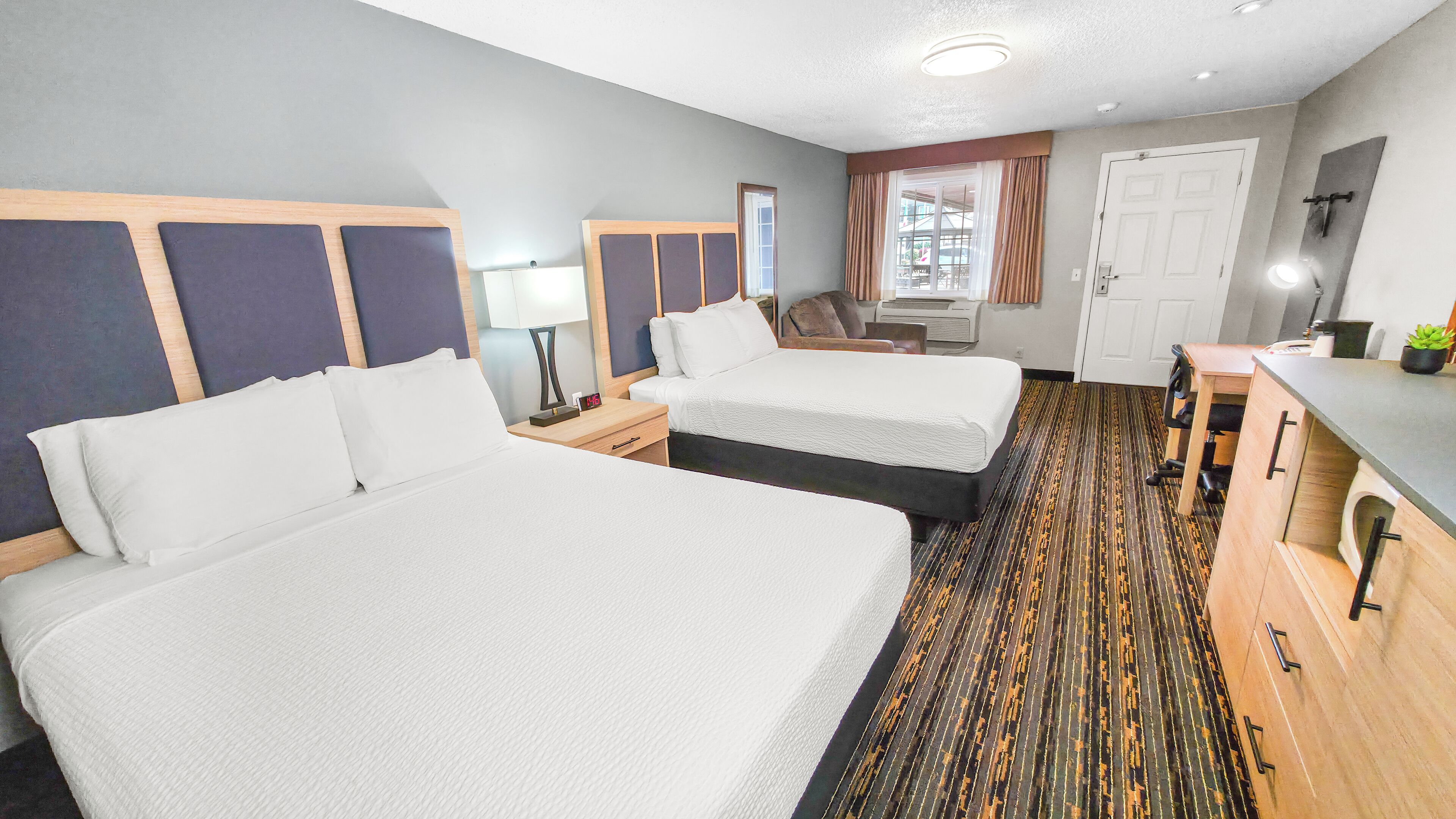 A hotel room with two beds, a desk, a chair, a window with curtains, and a door.