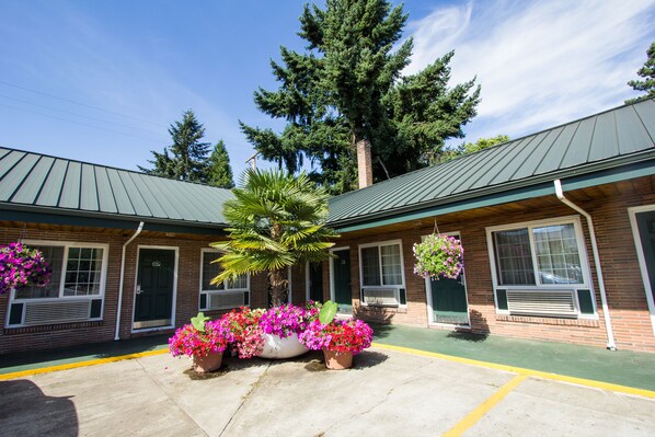 A building with a green roof, brick walls, and large windows. There are potted plants with pink flowers in front of the building.