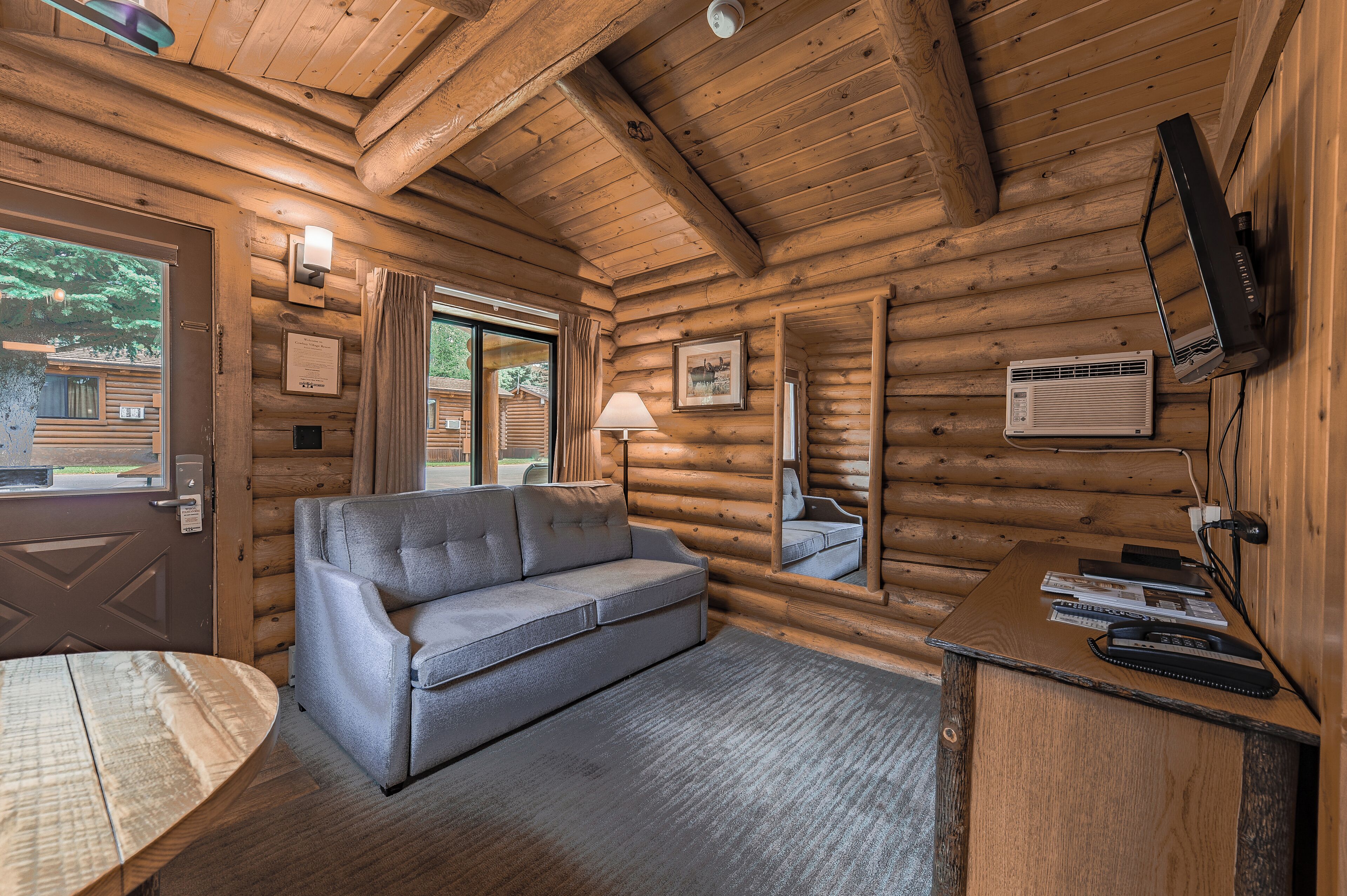 A log cabin interior with a sofa, a table, a TV mounted on the wall, and a window with curtains.