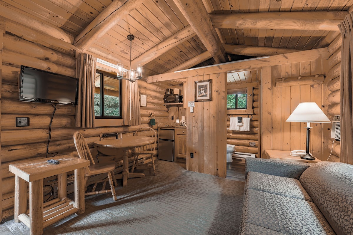 Interno di una baita di legno con zona pranzo, divano, TV a parete e angolo cottura.
