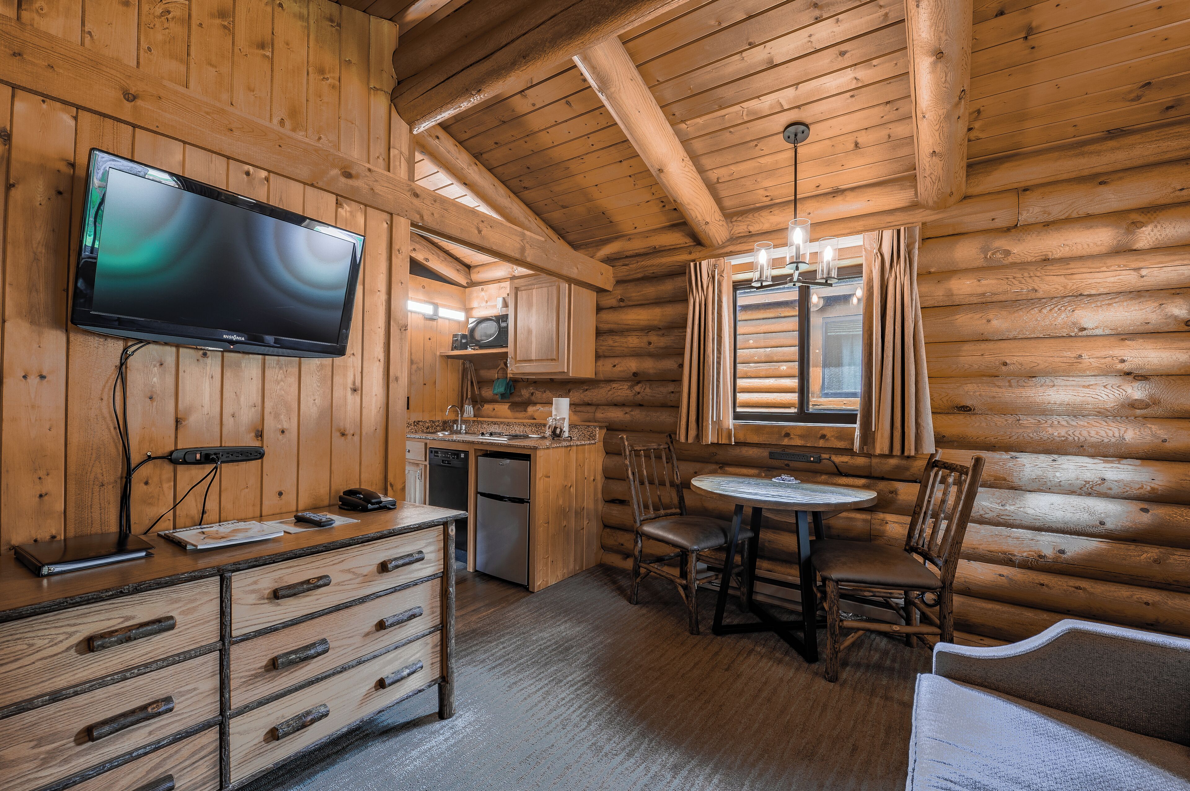 A log cabin interior with a wooden dining table, chairs, a television mounted on the wall, a kitchen area with a refrigerator and microwave, and a window with curtains.