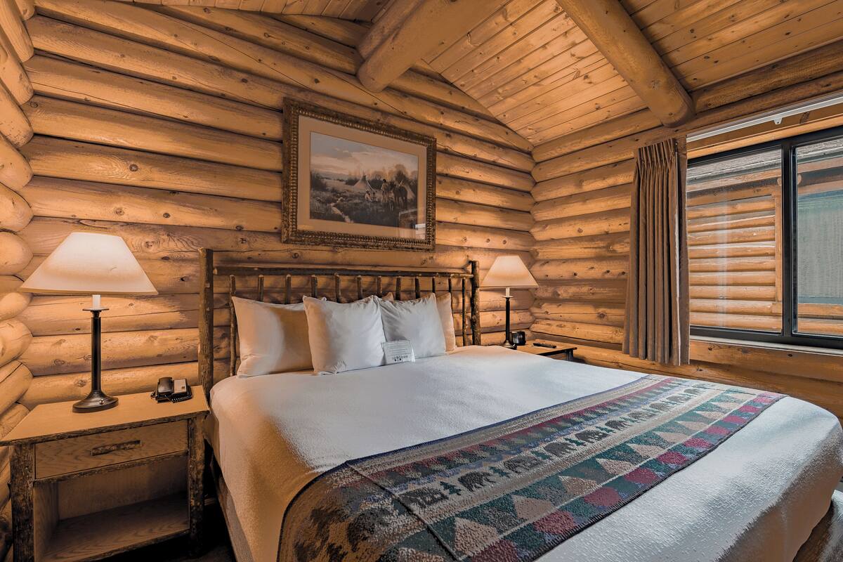 Camera in stile baita con una testiera in legno, due abat-jour, un letto con lenzuola bianche, un quadro appeso alla parete e una finestra con tende.