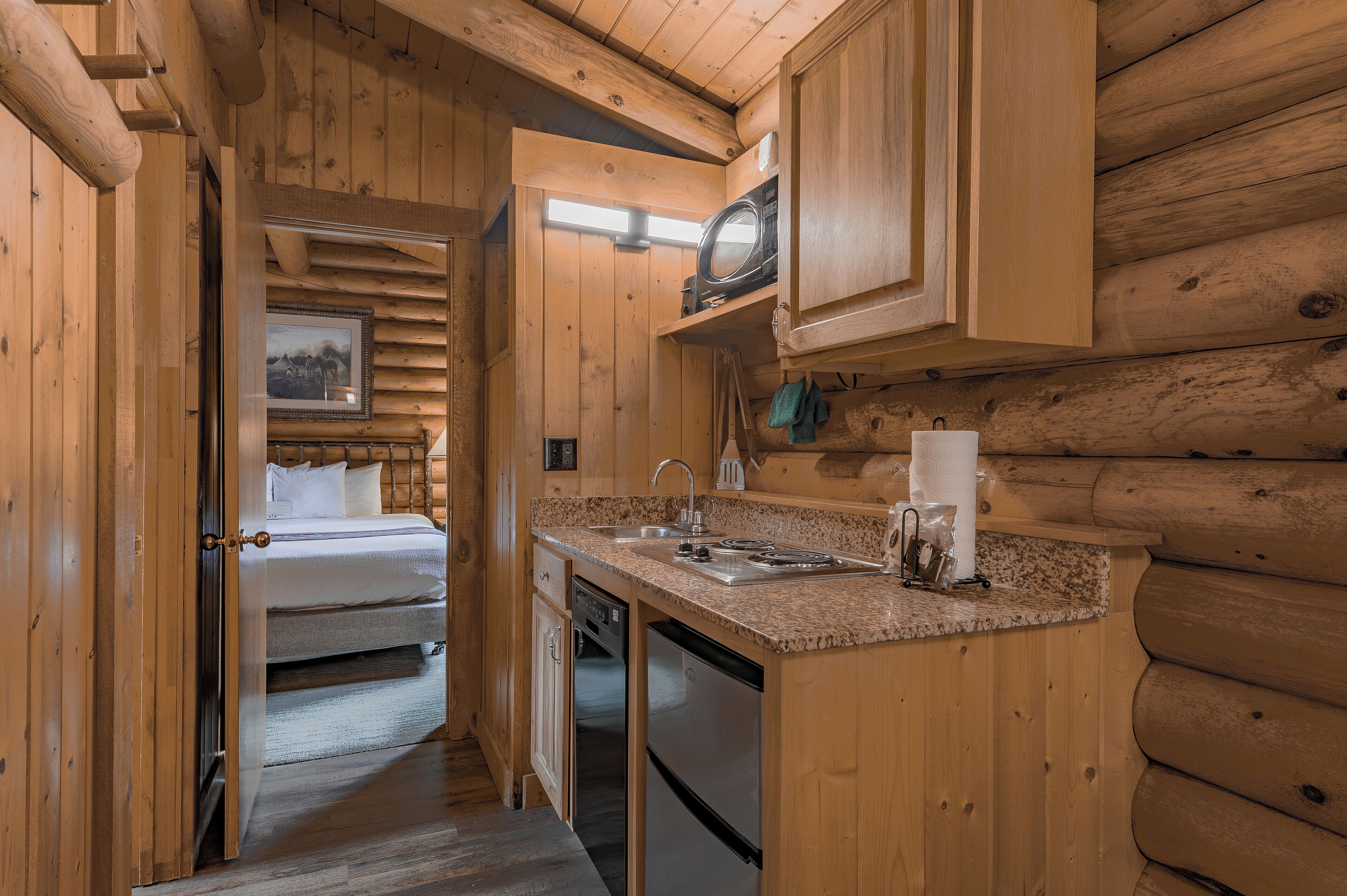 A log cabin interior with a kitchen area, a bed, and a bathroom.