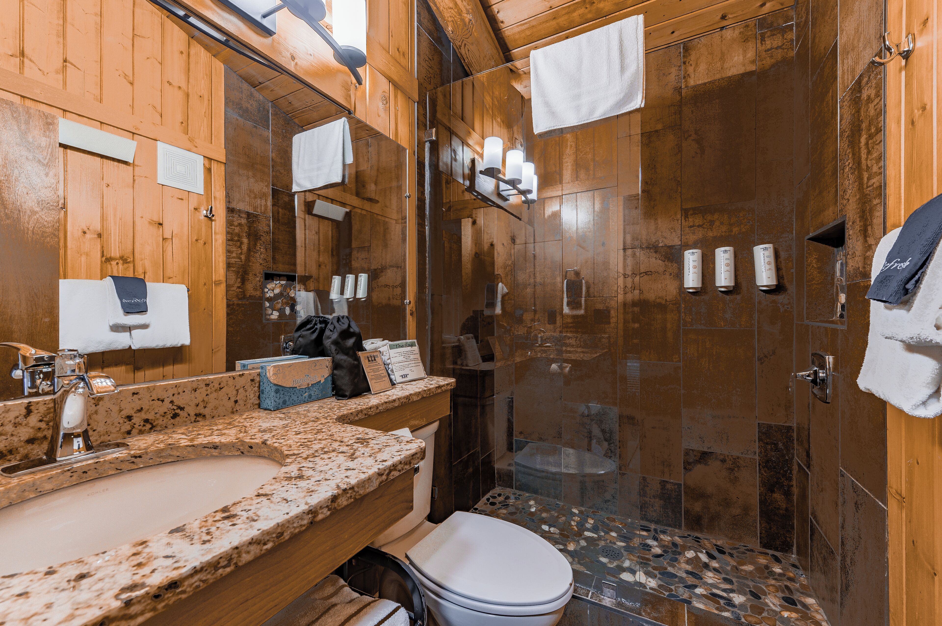 Een houten badkamer met een wastafelblad van graniet, een toilet en een douche met glazen wanden.