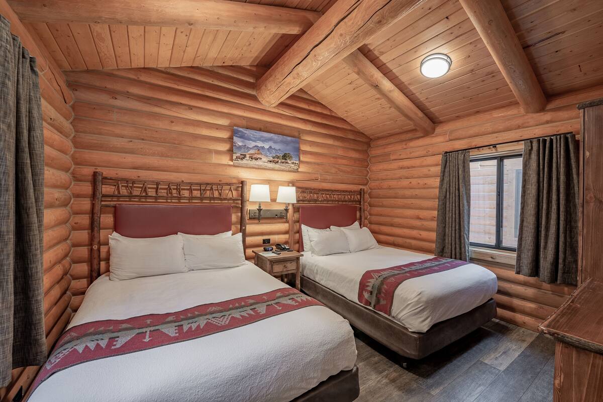 Una stanza di una baita di legno con due letti, pareti in legno, un quadro di montagne e una finestra con tende.