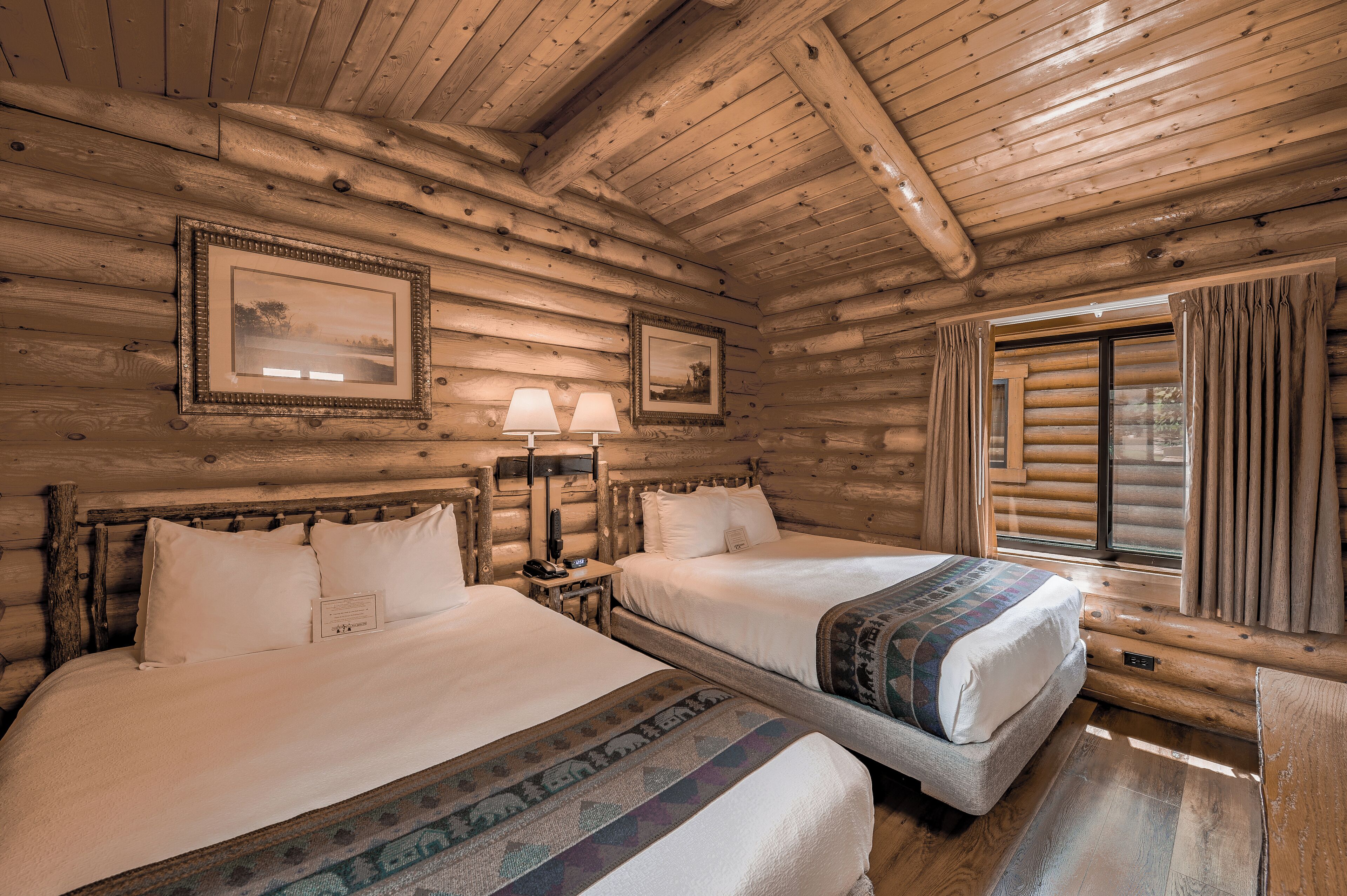 A log cabin room with two beds, framed pictures on the walls, and a window with curtains.
