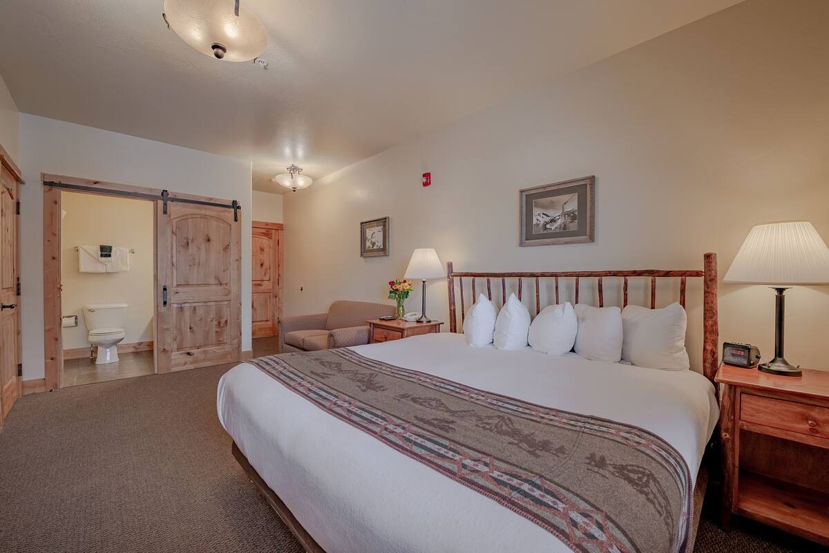 Una camera da letto con un grande letto, una testiera in legno, un comodino con una lampada, un quadro incorniciato appeso al muro e una porta che dà su un'altra stanza.