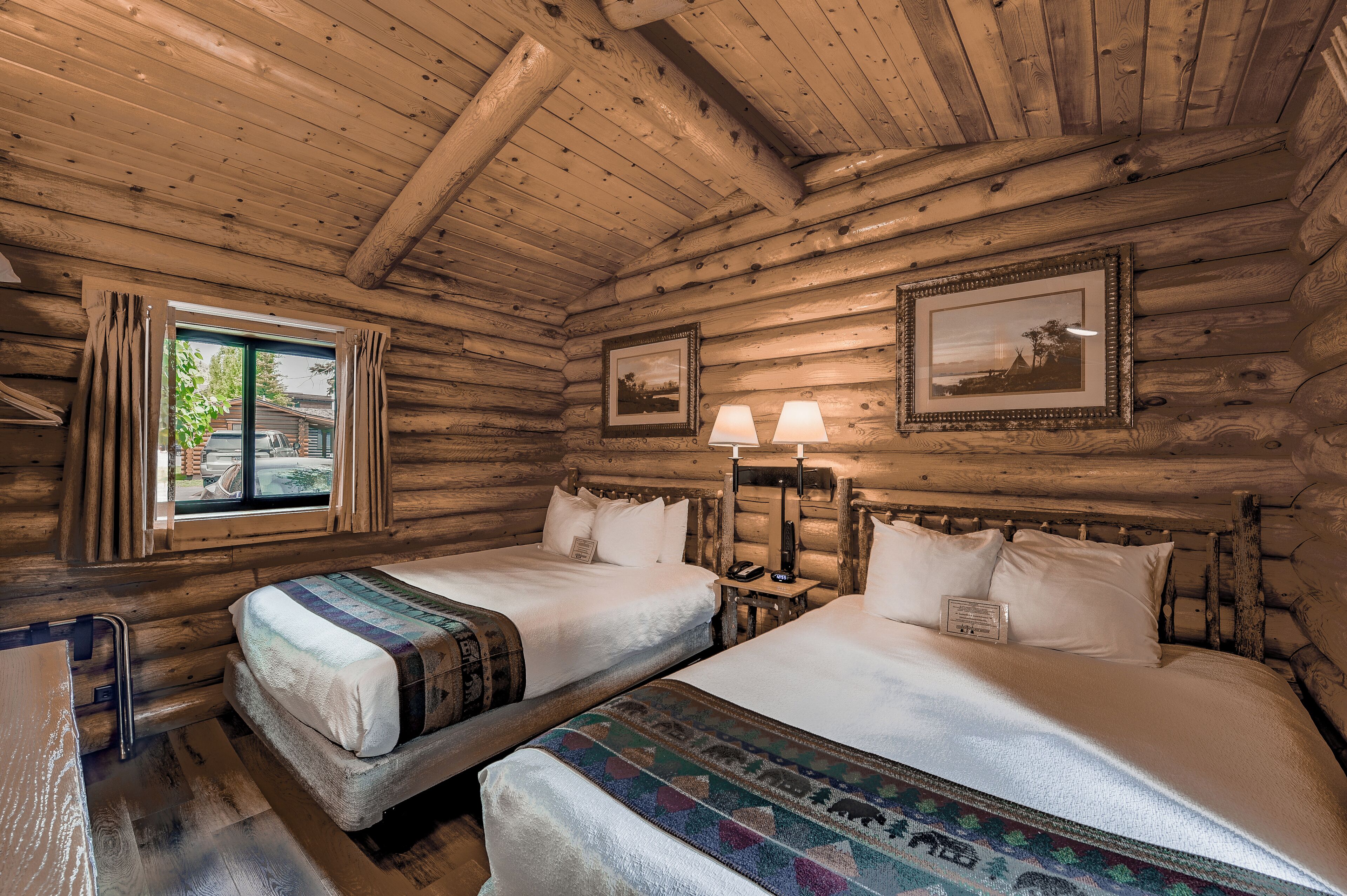 A log cabin room with two beds, a window with curtains, a framed picture on the wall, and a small table with a lamp.