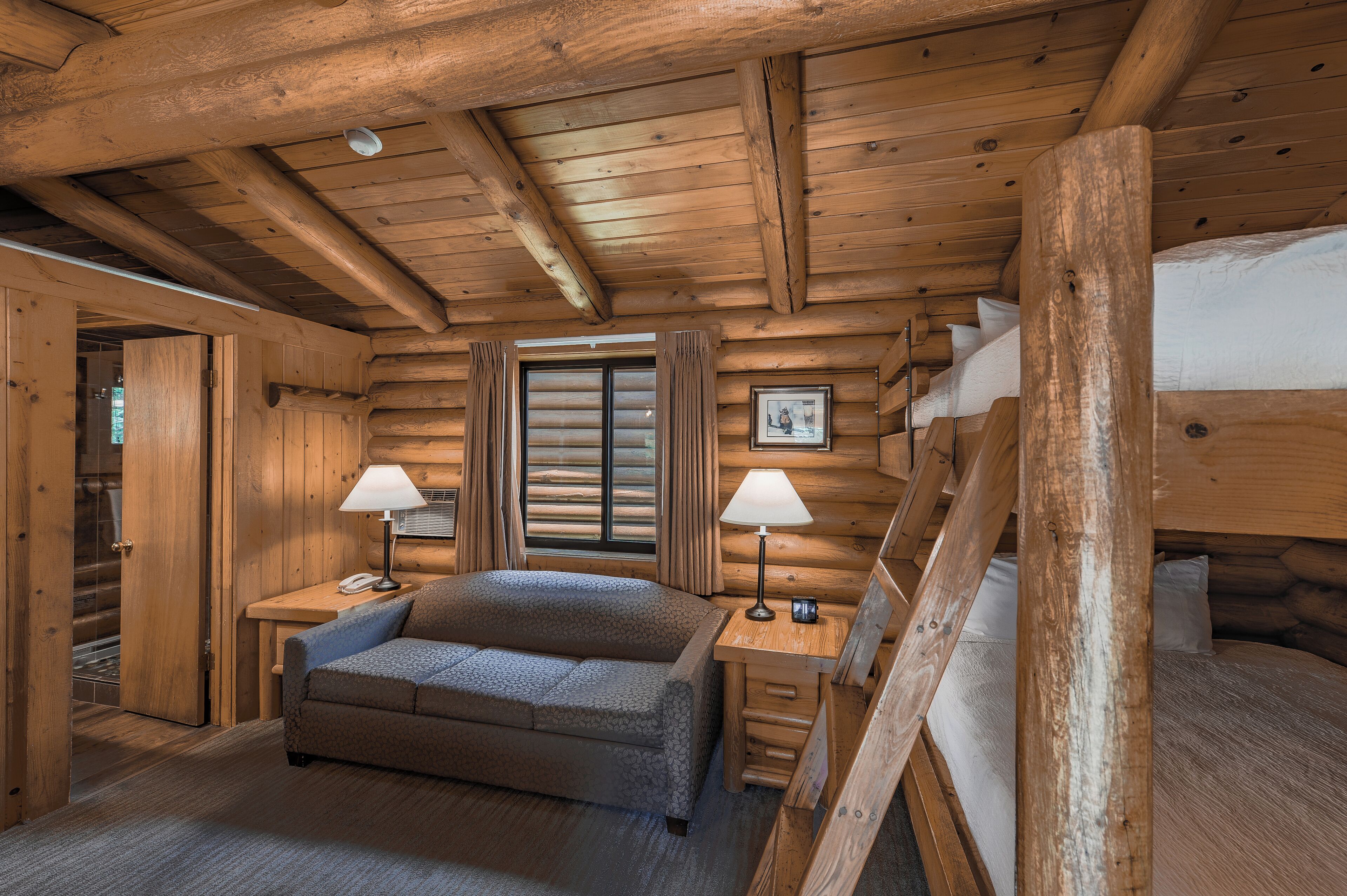 A log cabin bedroom with a bed, two bedside tables with lamps, a sofa, and a window with curtains.