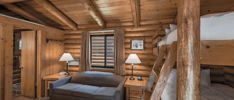 A log cabin bedroom with a bed, two bedside tables with lamps, a sofa, and a window with curtains.