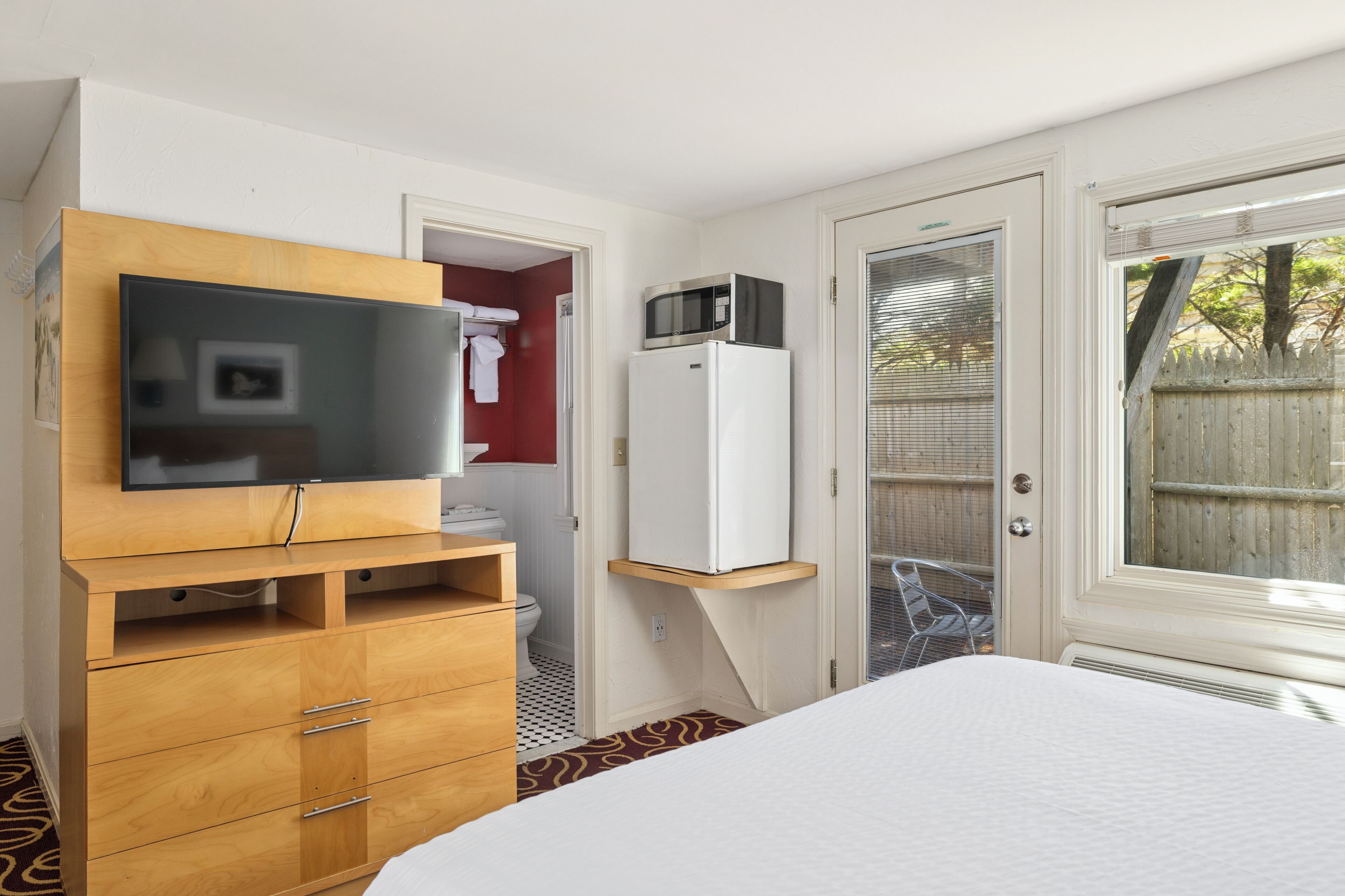 A bedroom with a bed, a television on a wooden dresser, a microwave on a white refrigerator, and a sliding glass door to the outside.