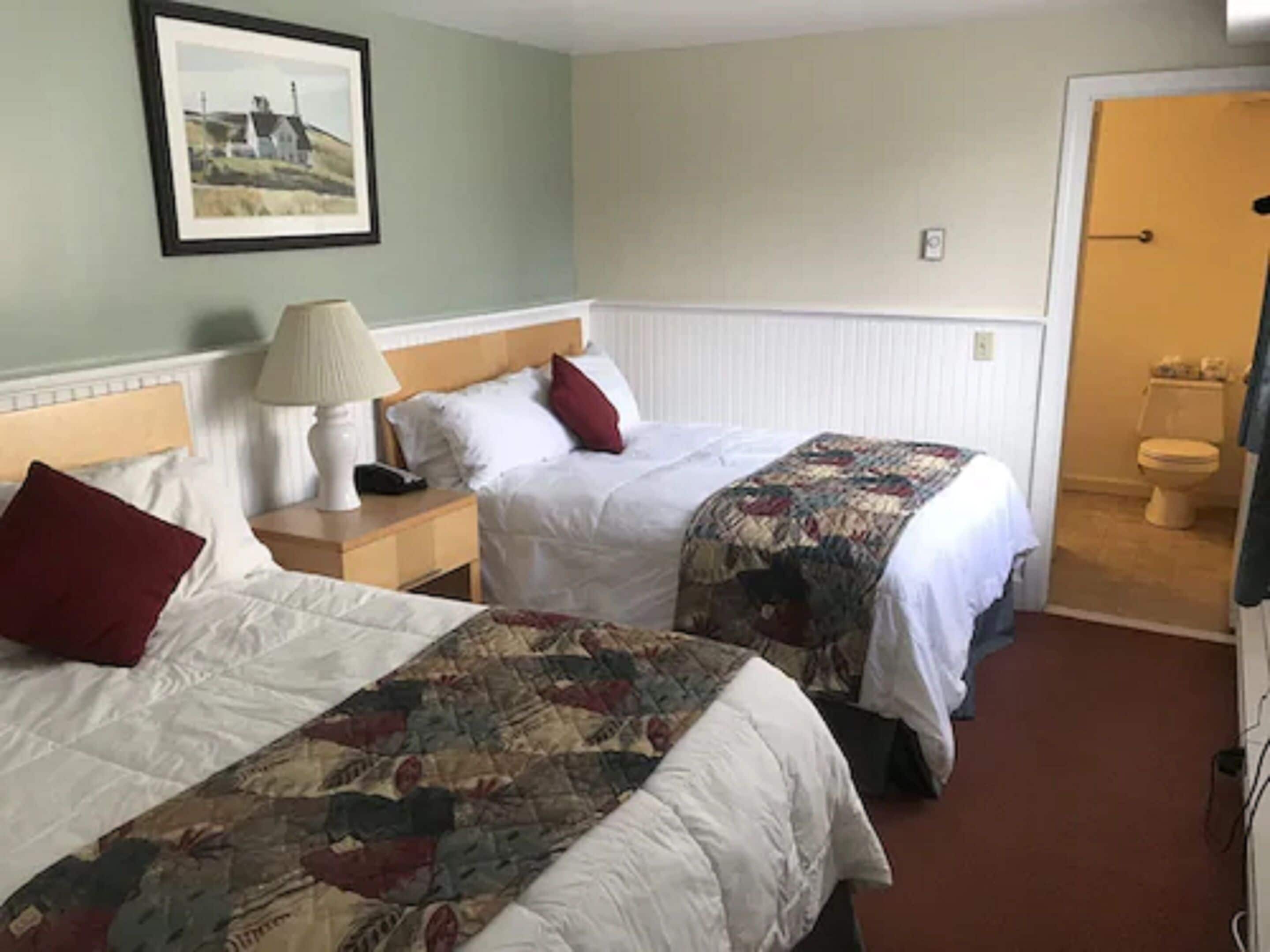 A hotel room with two beds, a nightstand with a lamp, a framed picture on the wall, and a view of the bathroom.