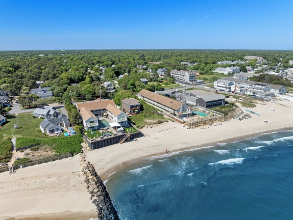 An aerial view of a coastal resort with a large building featuring a swimming pool, beach access, and a rocky shoreline. - The Corsair & Cross Rip Oceanfront Resort (Dennis Port)
